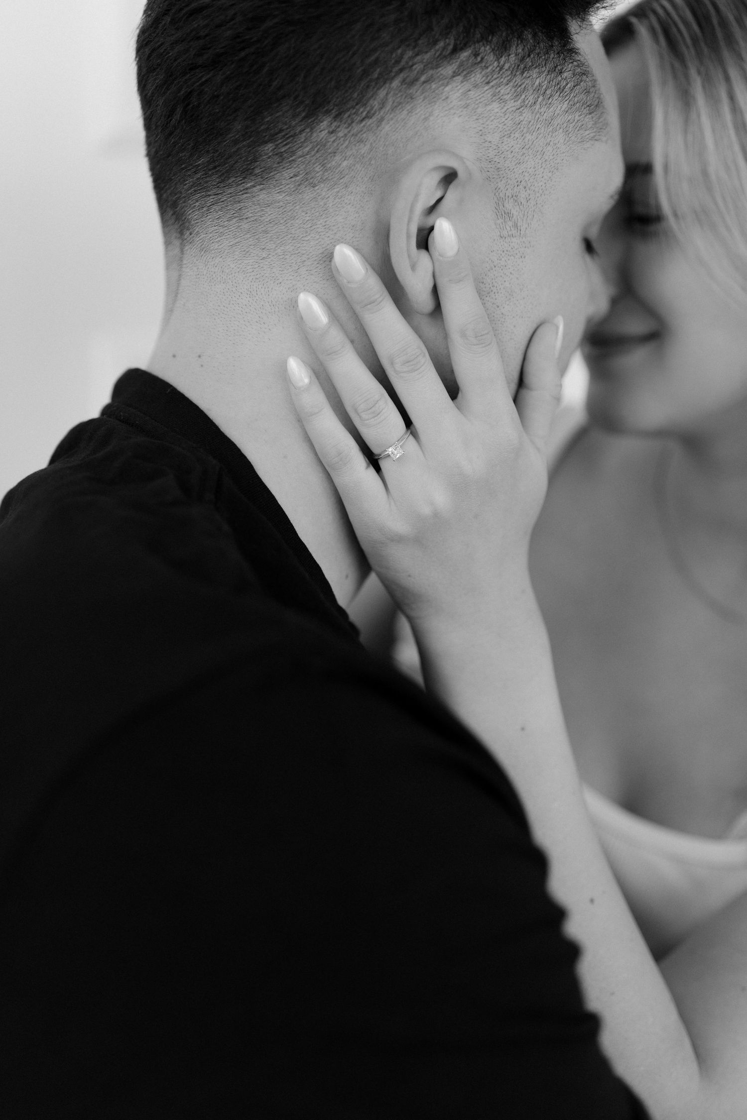 Black and white close-up of couple almost kissing, woman's hand with engagement ring tenderly cupping man's face.