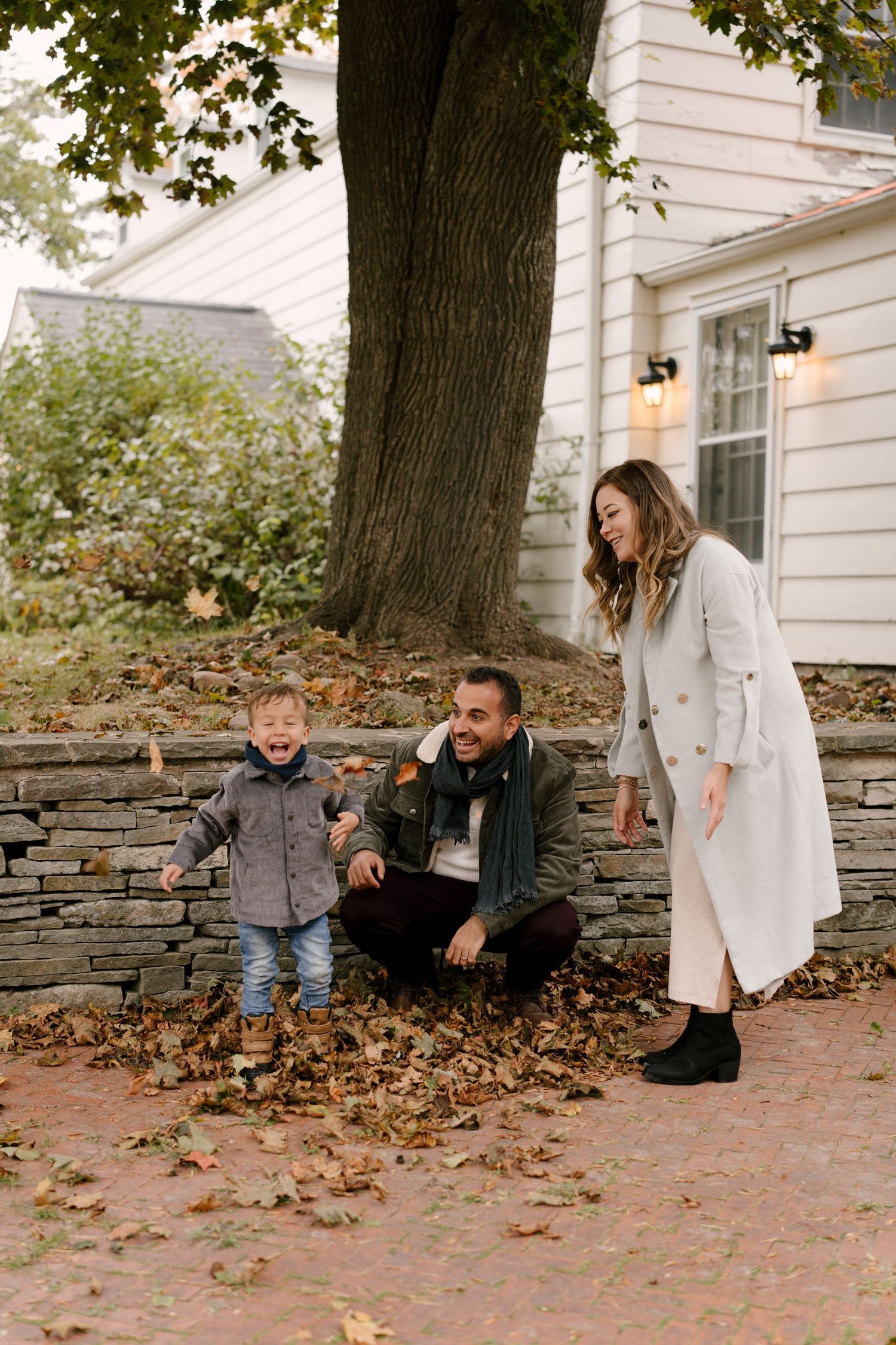 Joyful toddler laughing while playing in fall leaves with parents outside historic home.