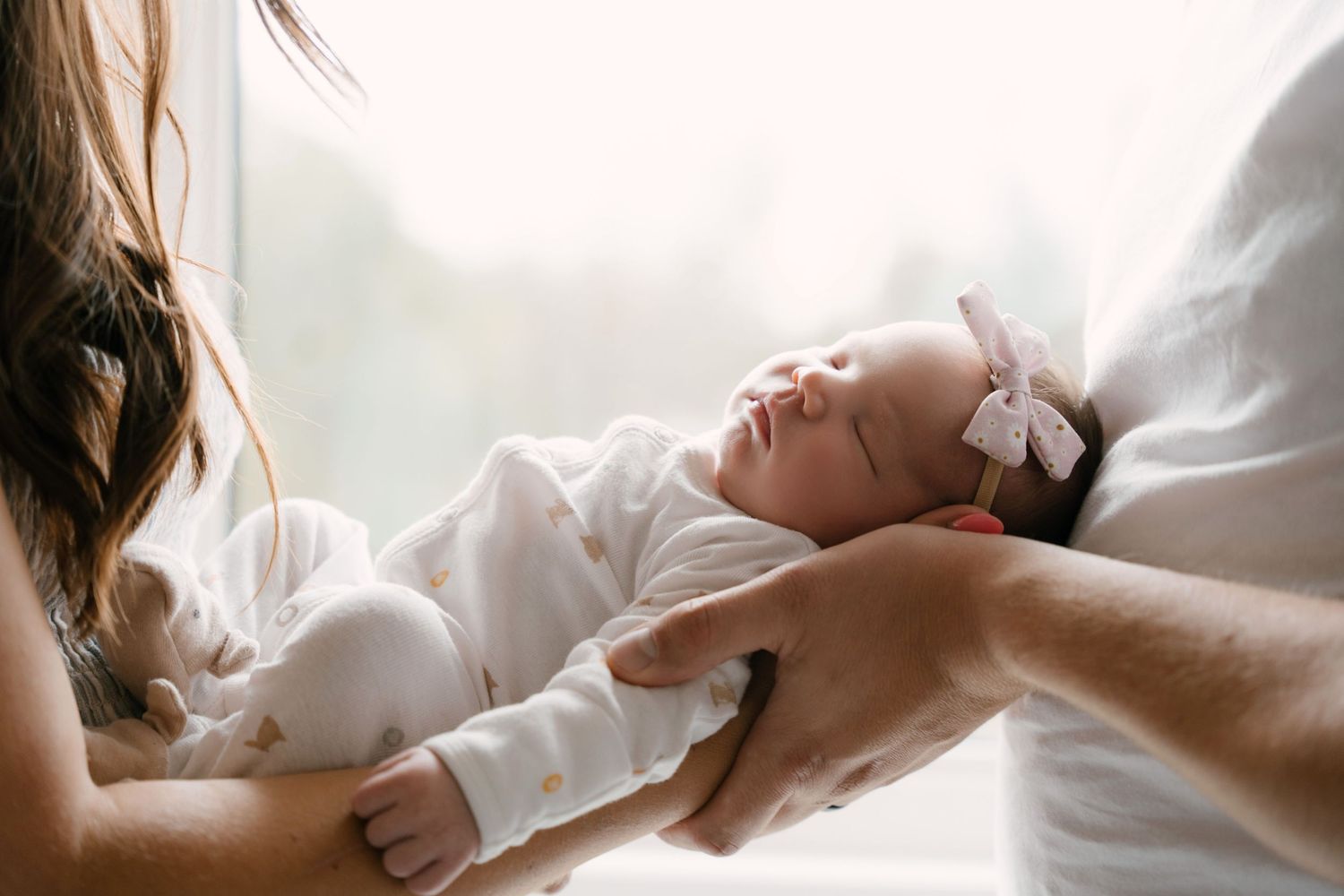 Newborn baby sleeping peacefully in parents arms wearing white outfit and pink bow headband.