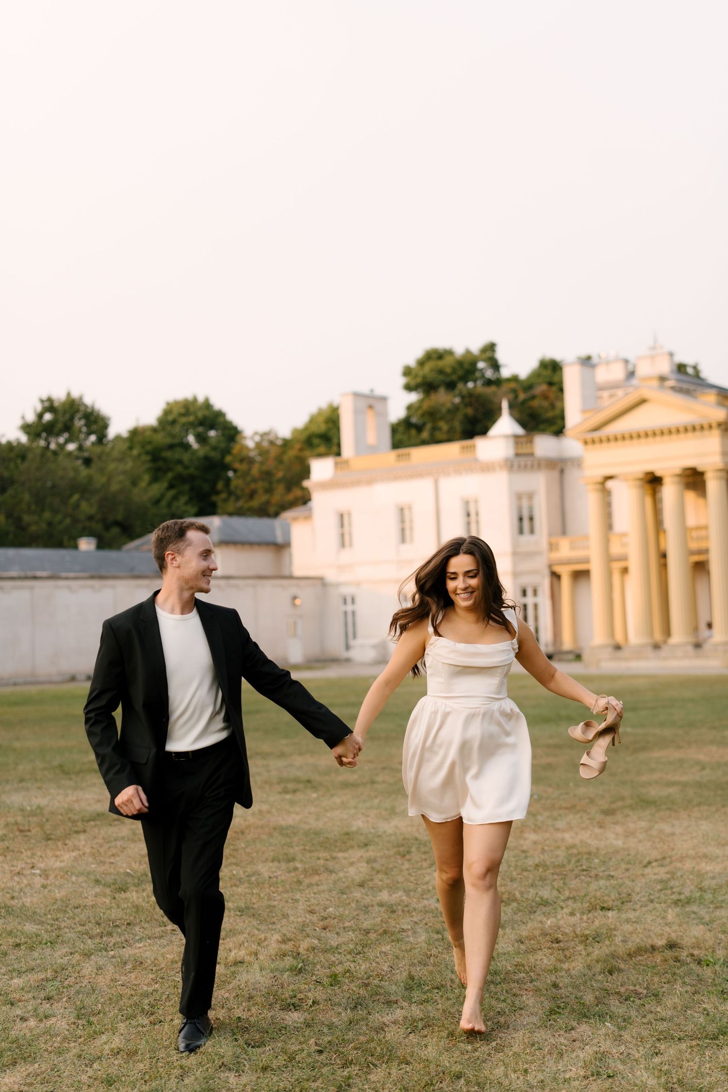 Joyful couple holding hands and running barefoot across a lawn near a grand estate.