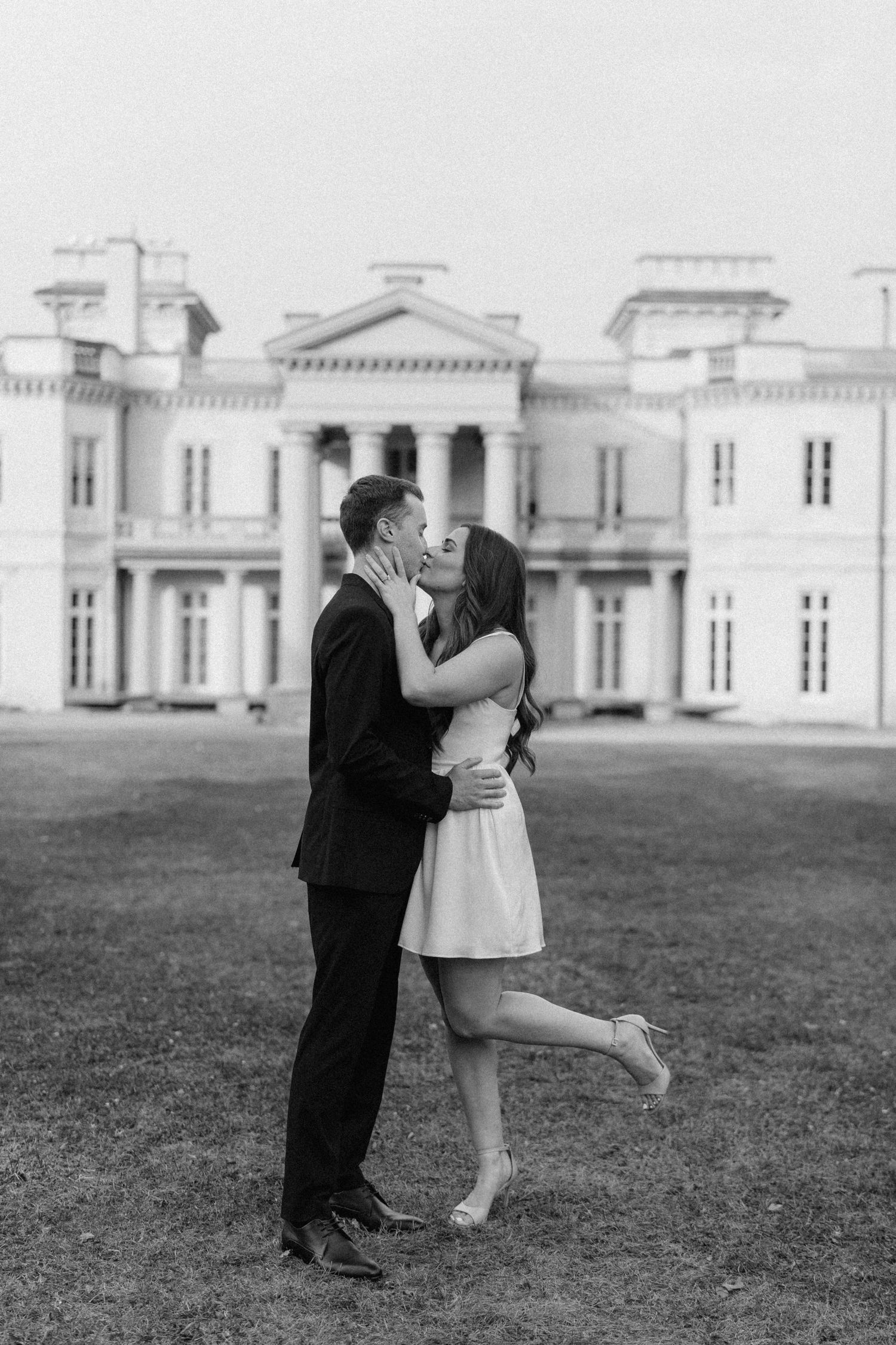Black and white photo of couple kissing on lawn in front of grand estate.