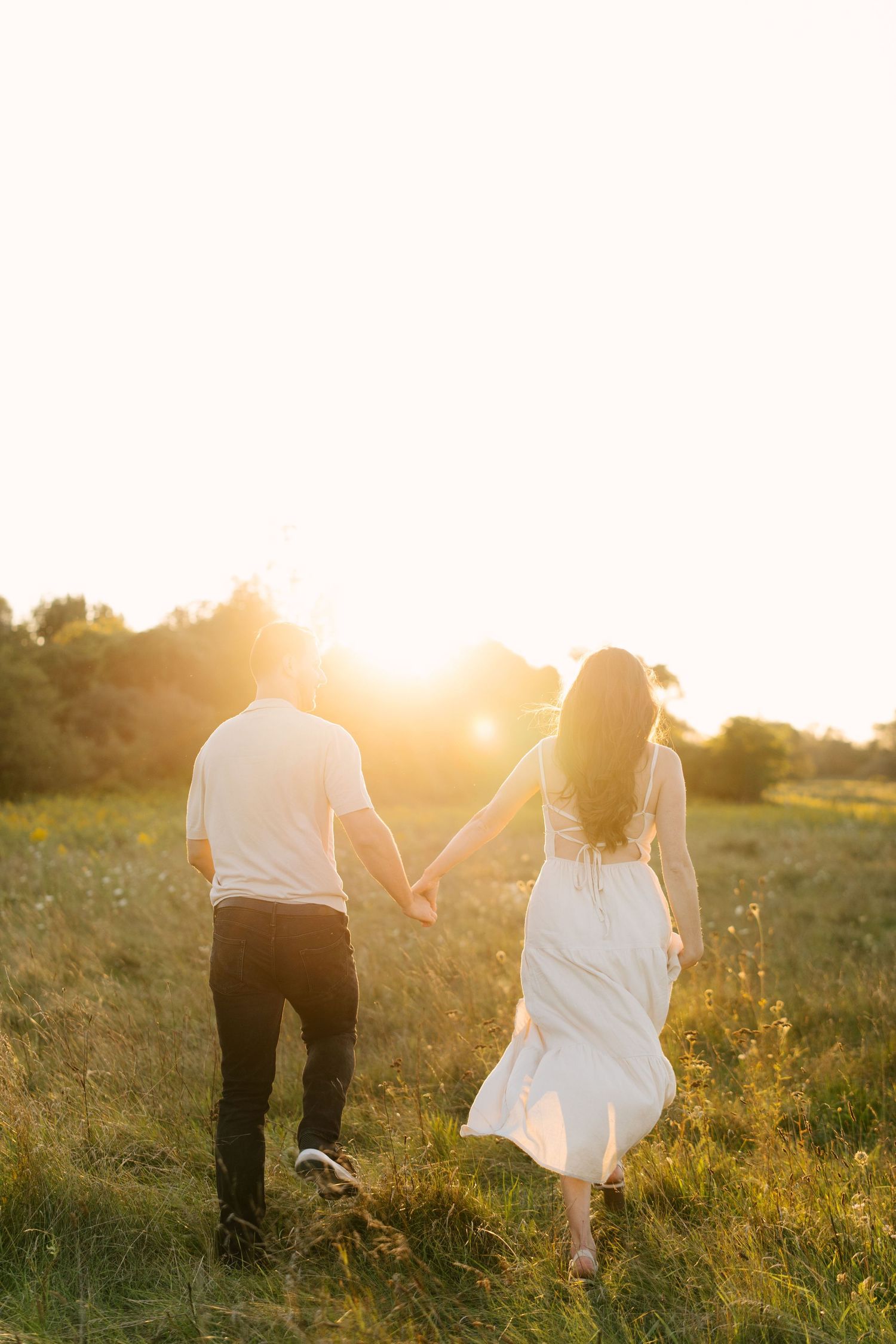 Couple walking hand in hand toward glowing sunset in lush green field, woman in flowing white dress.