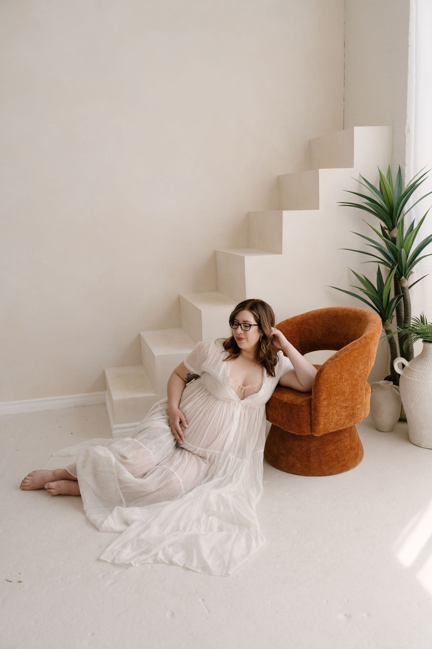 Pregnant woman in sheer white gown sitting on floor beside orange chair and staircase in bright studio.