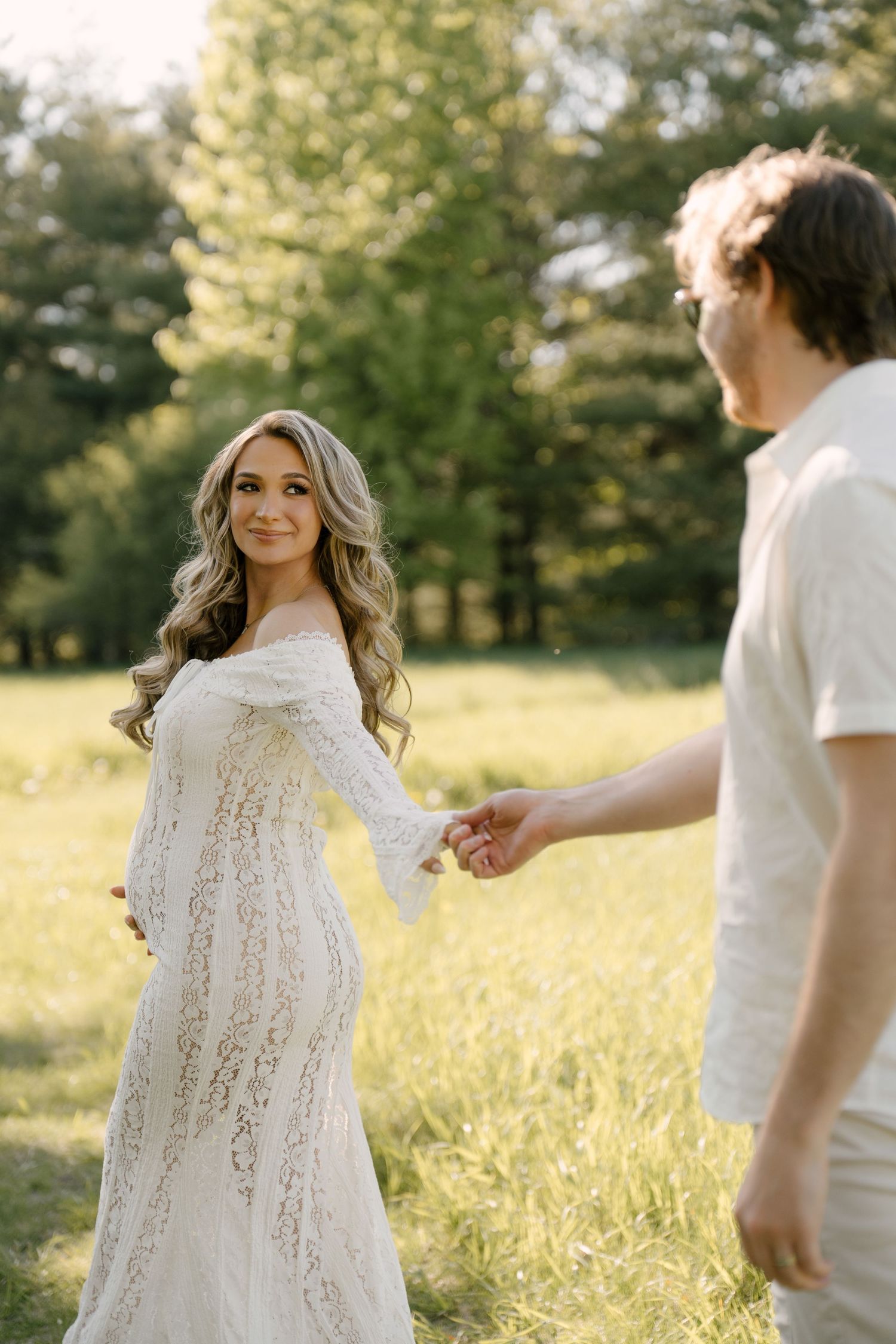 Smiling pregnant woman in white lace gown holds partner's hand in a sunlit green meadow.