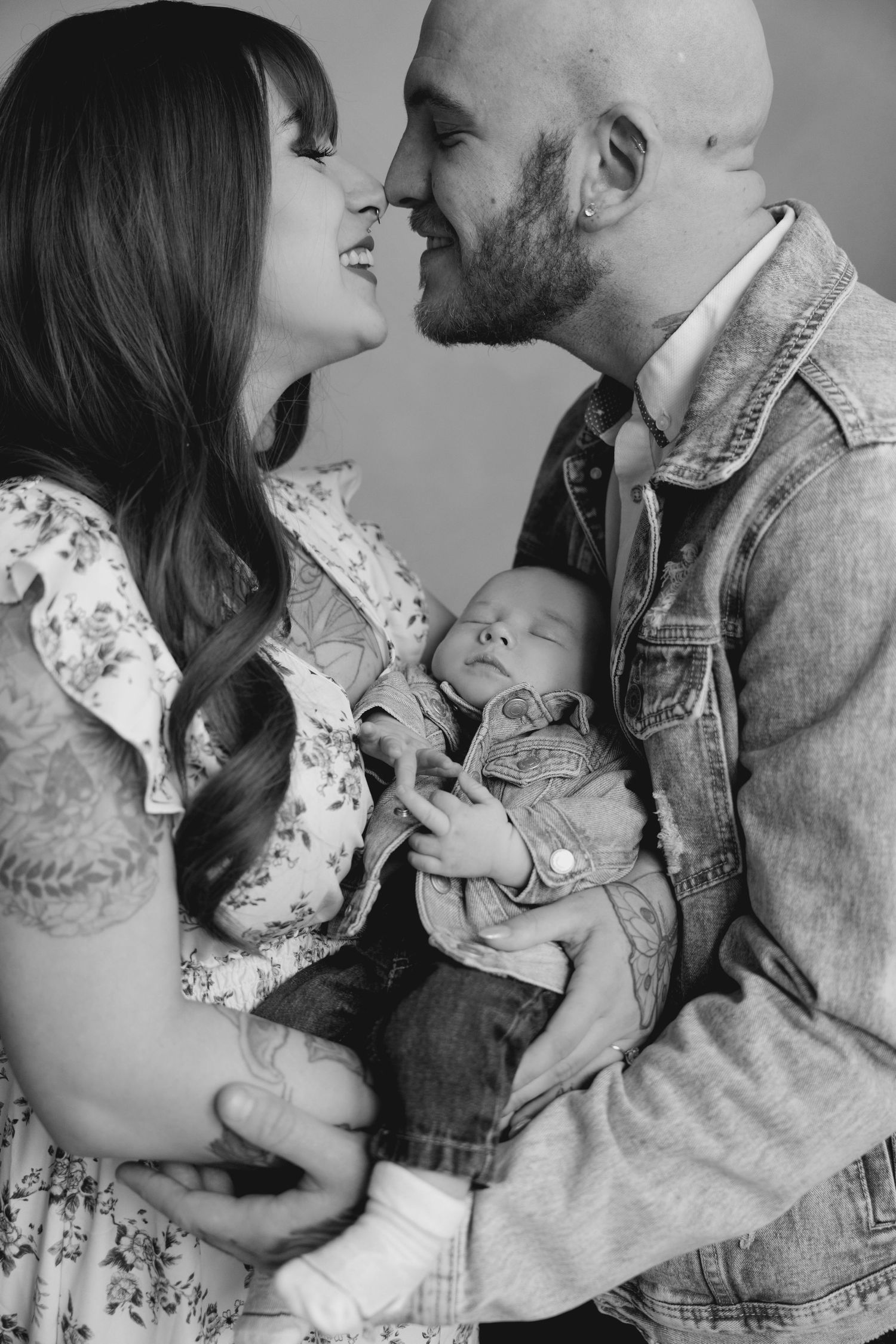 Black and white photo of tattooed parents smiling at each other while holding their newborn baby.