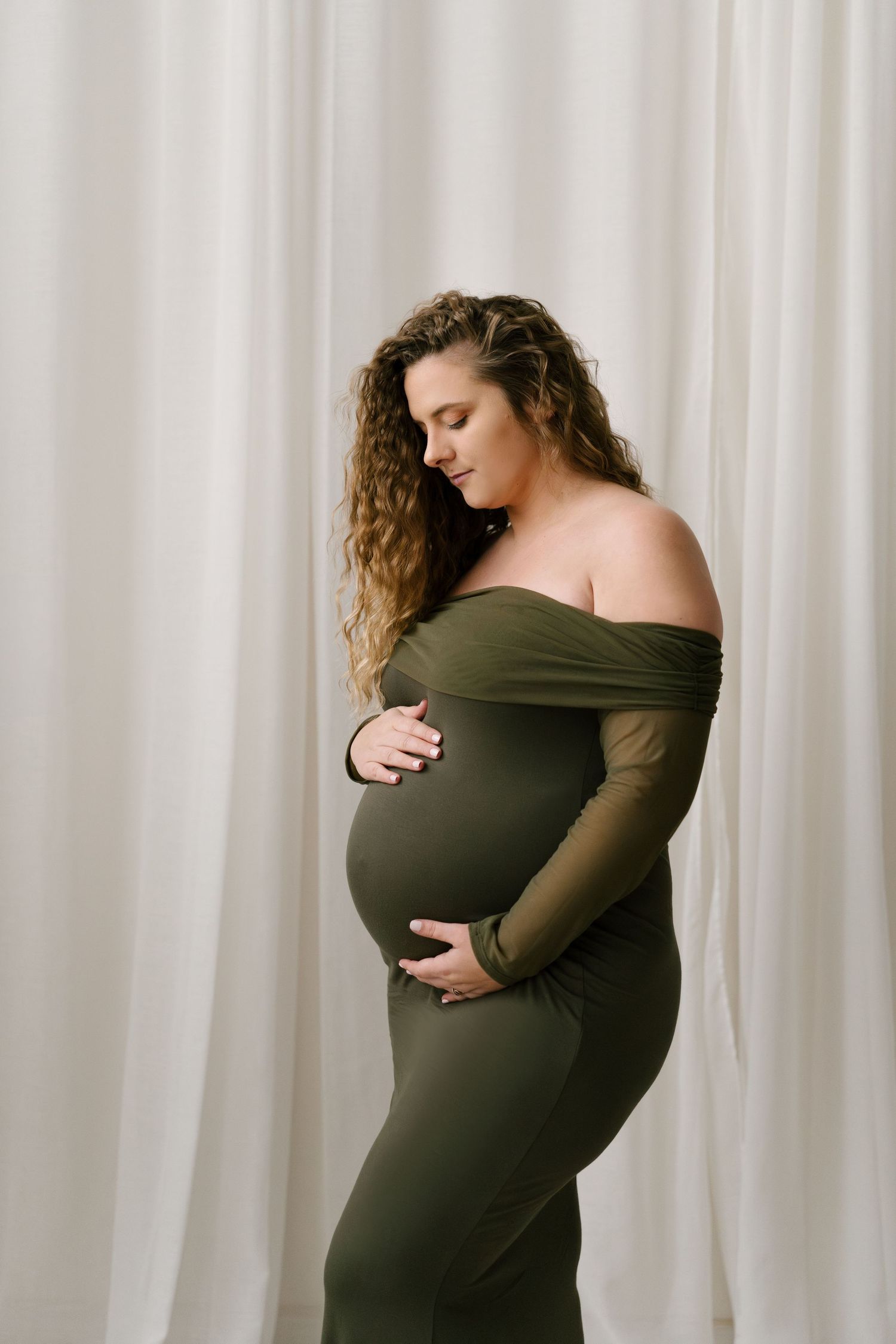 Curly-haired pregnant woman in green off-shoulder dress gently holding baby bump indoors.