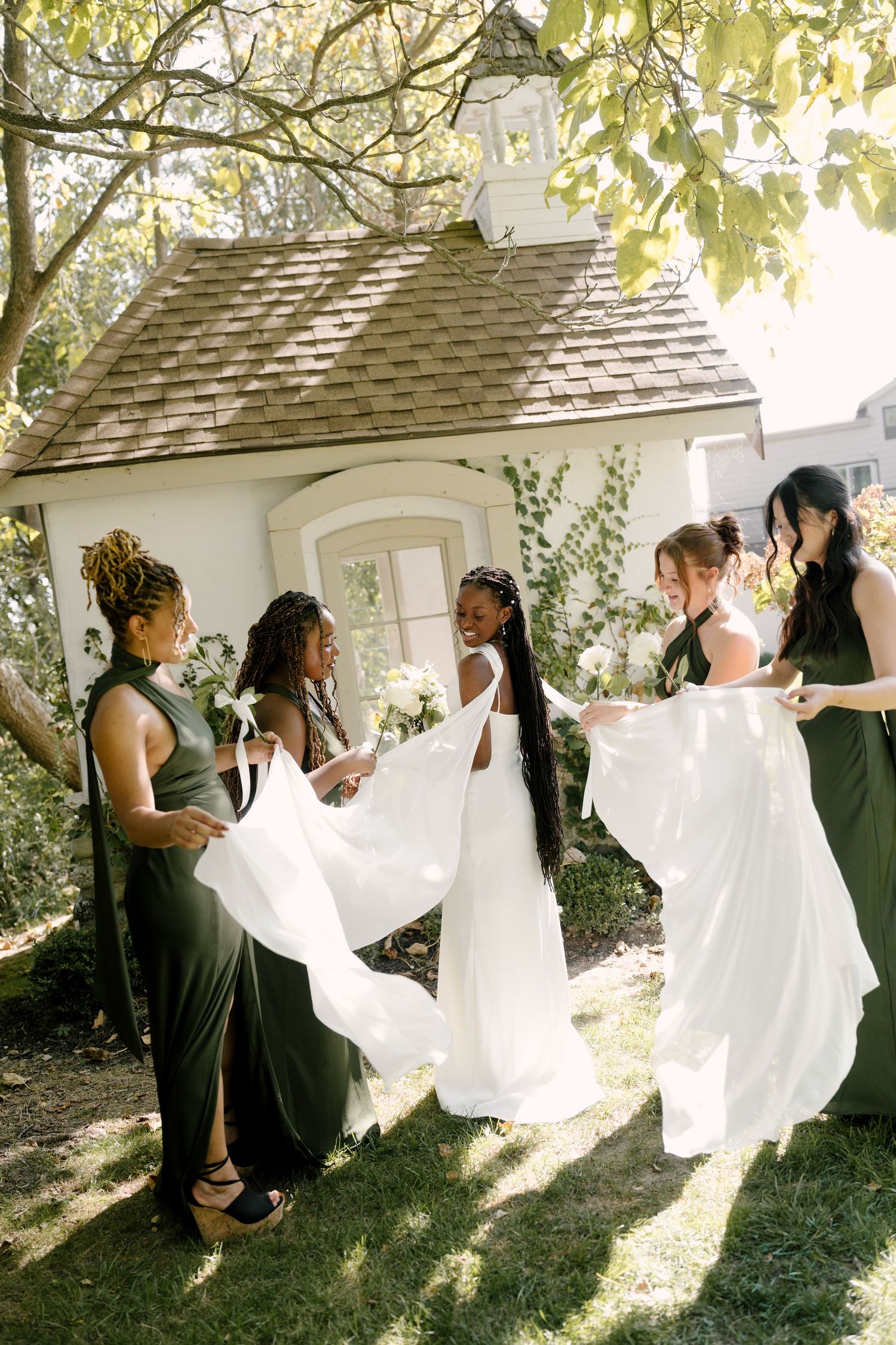 Black bride in white gown laughing with bridesmaids in olive green dresses near a garden chapel at Wren House.