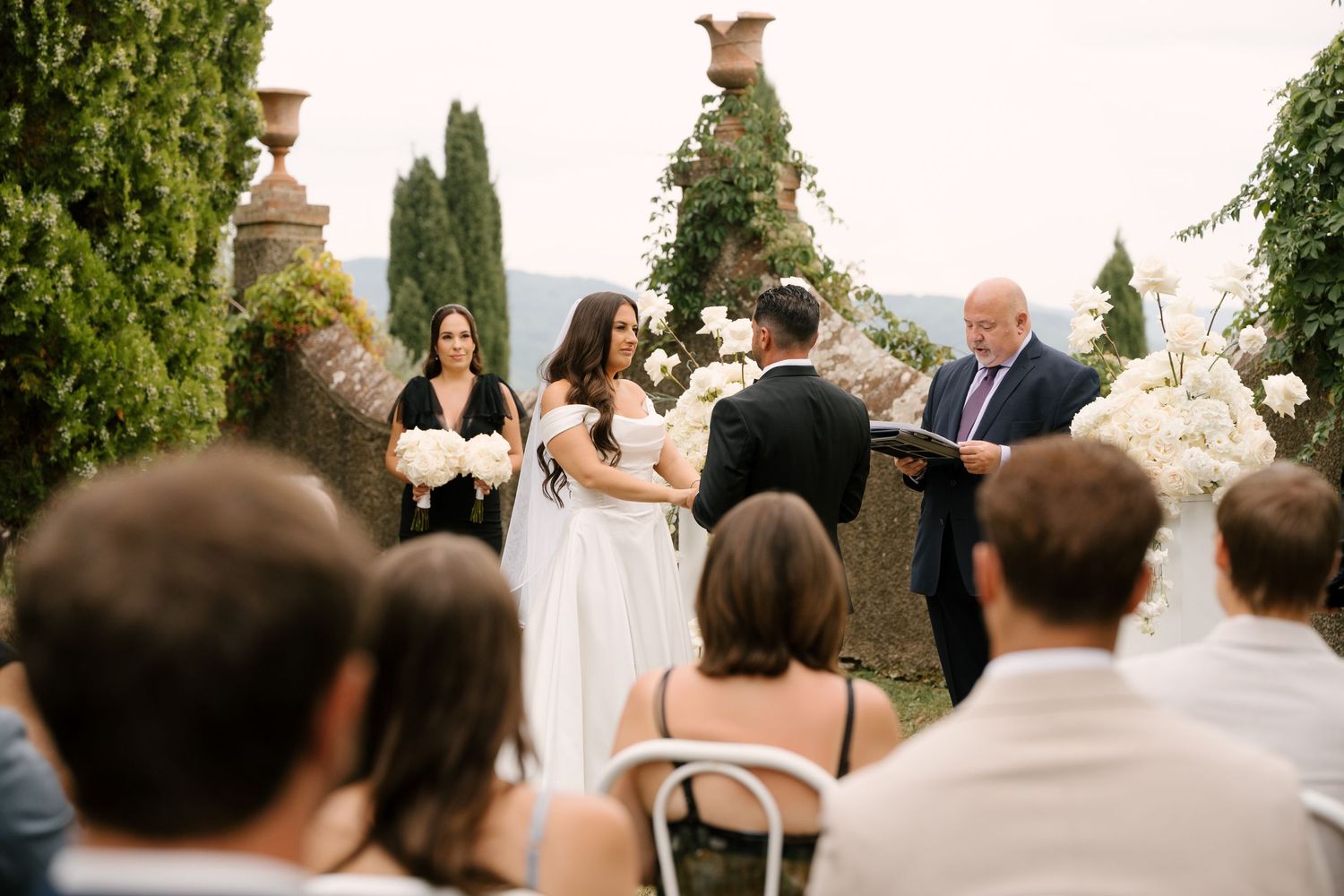 Bride and groom exchange vows at elegant outdoor wedding ceremony with white floral arrangements and cypress trees.
