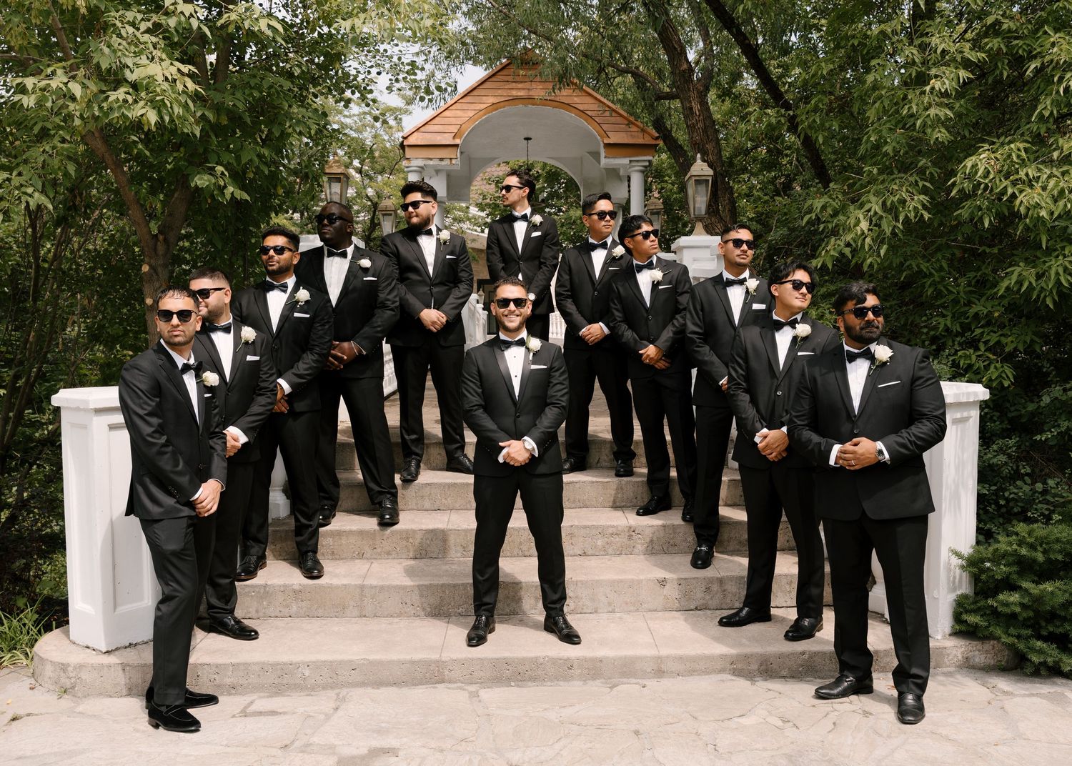 Groom and groomsmen in matching black tuxedos with bow ties pose confidently on outdoor stone staircase.