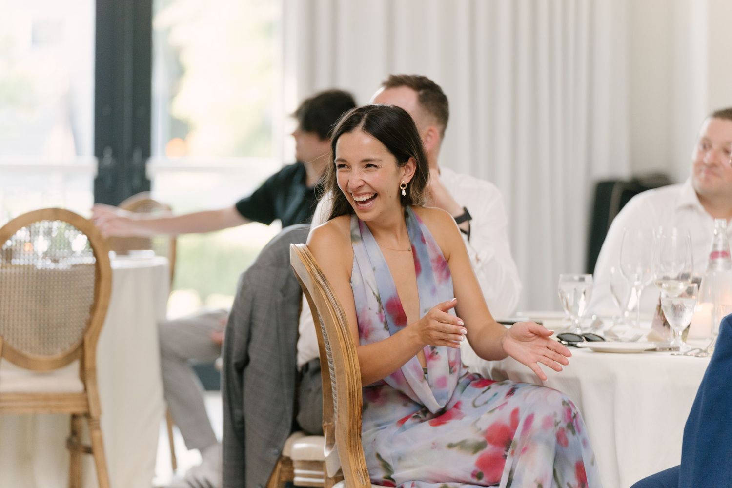 Smiling wedding guest in floral dress laughing at reception dinner table with guests.