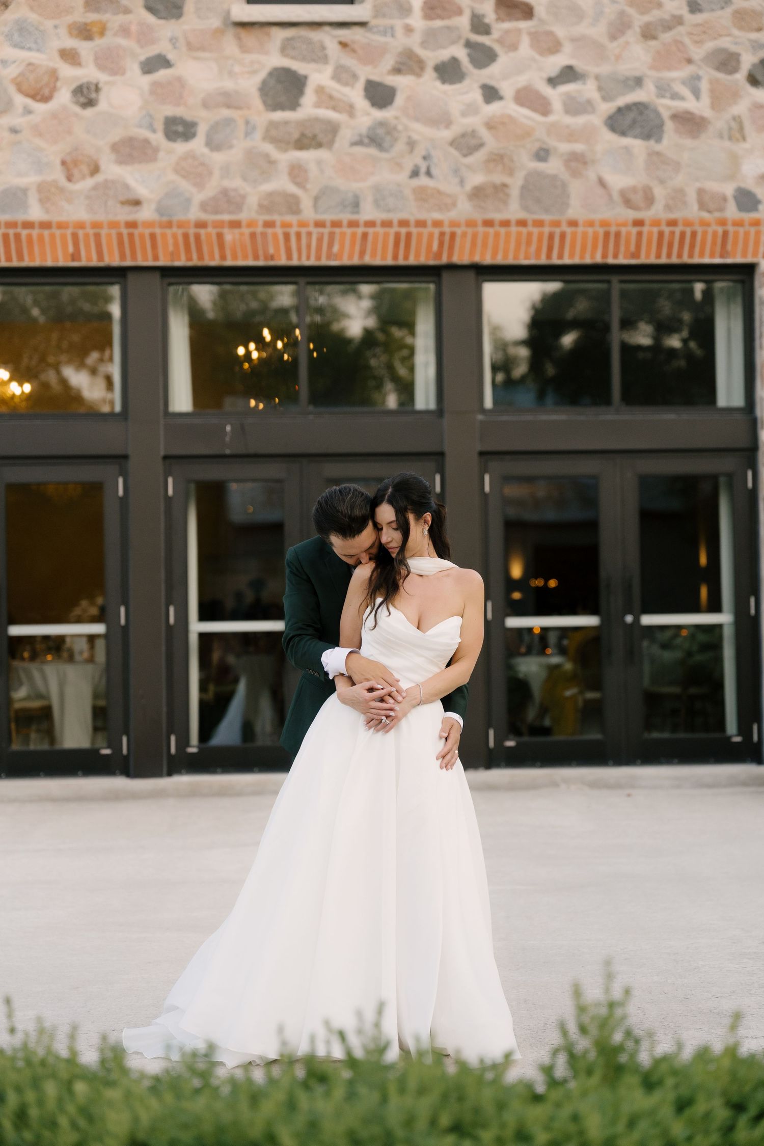 Groom embracing bride from behind in front of a stone venue with large glass doors at golden hour at Roseville Estates wedding venue.