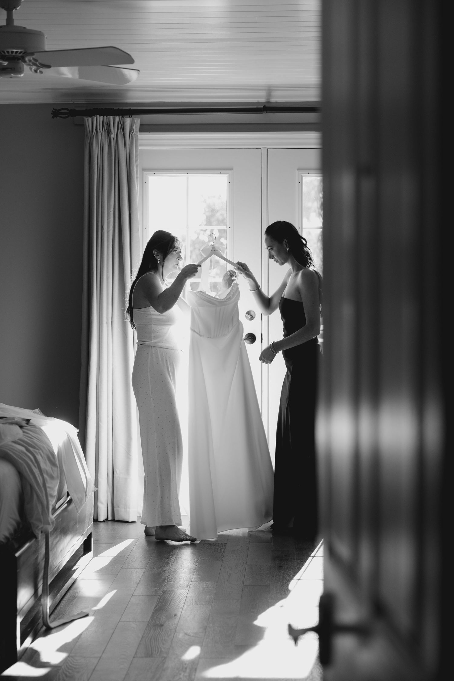 Two women hold up white wedding dress near sunlit French doors in black and white photo.