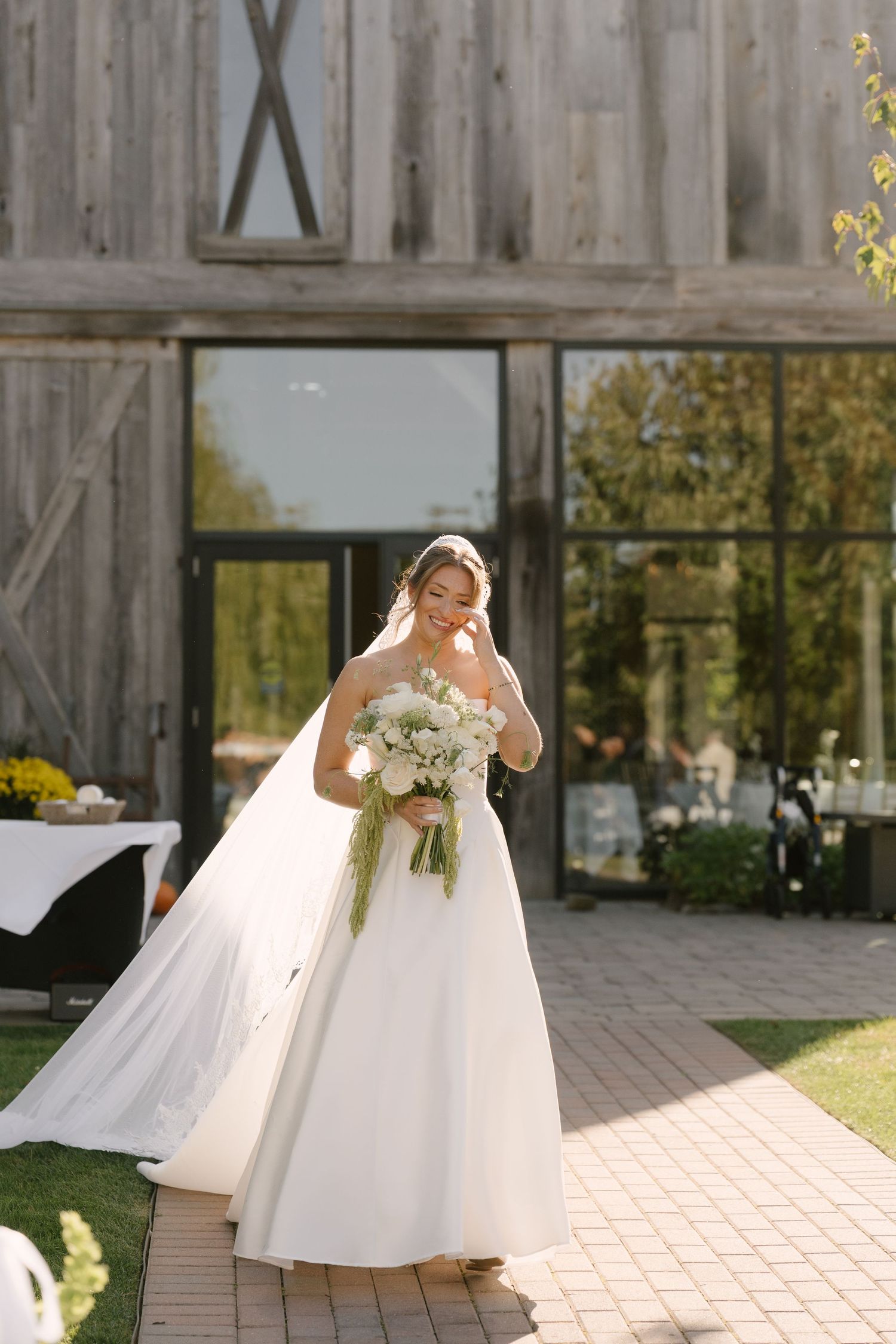 Bride in white ball gown holding floral bouquet stands before rustic barn venue on sunny wedding day at Pillar and Post.