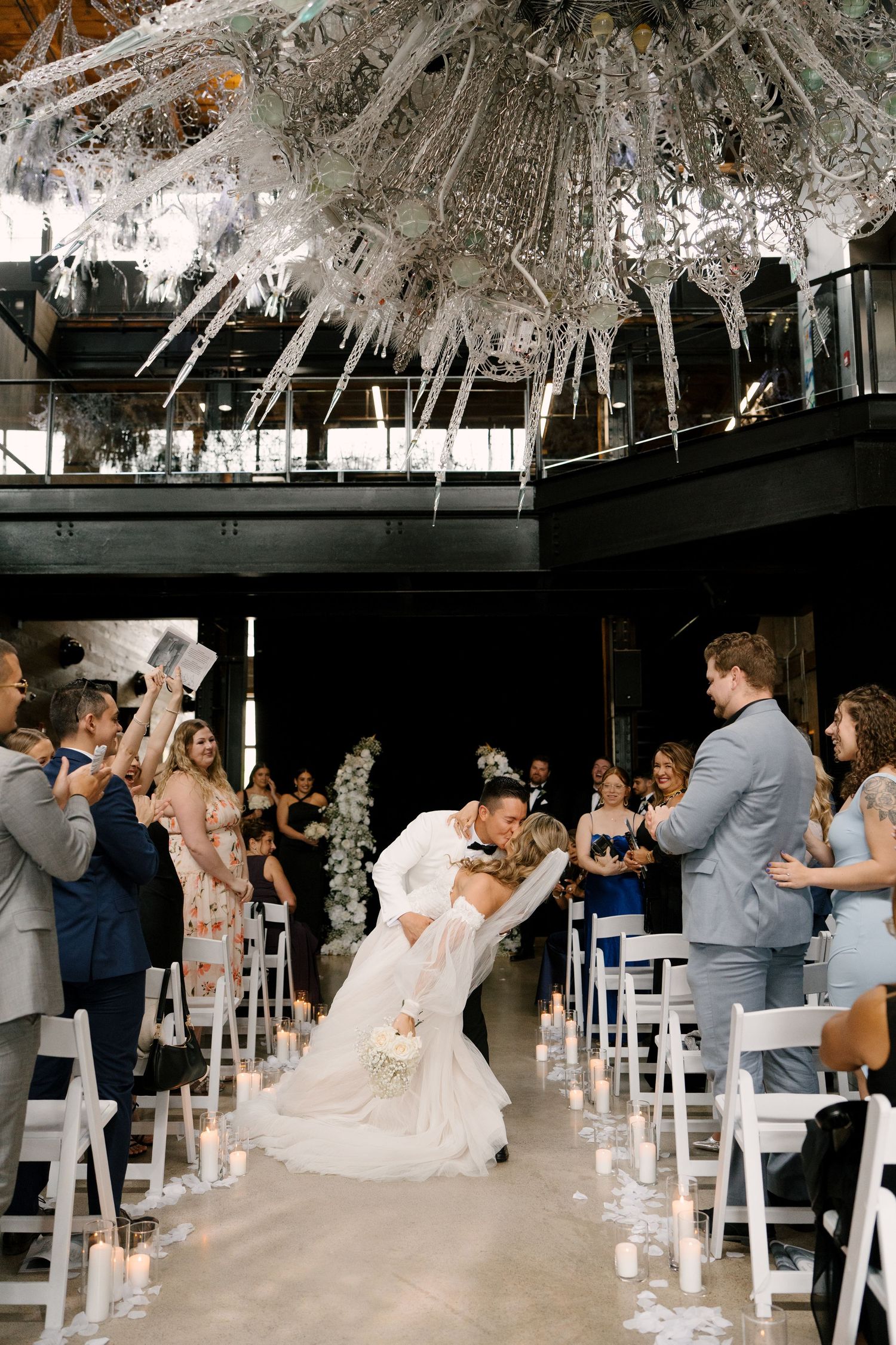 Groom dipping bride for a kiss in the aisle after wedding ceremony beneath a dramatic white sculptural chandelier at Tapestry Hall venue.