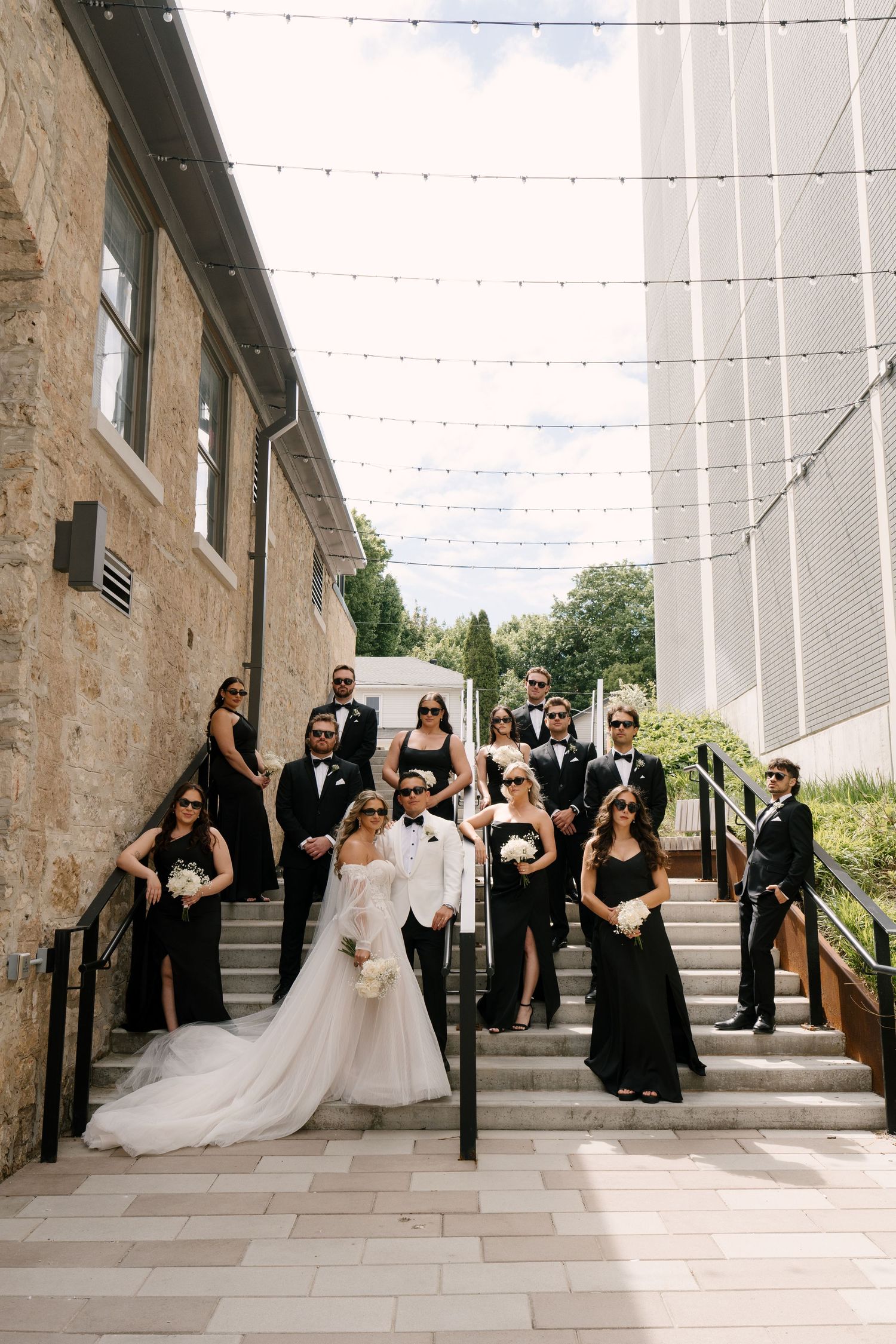 Wedding party in black and white attire posing on outdoor steps with string lights overhead at Tapestry Hall wedding venue.