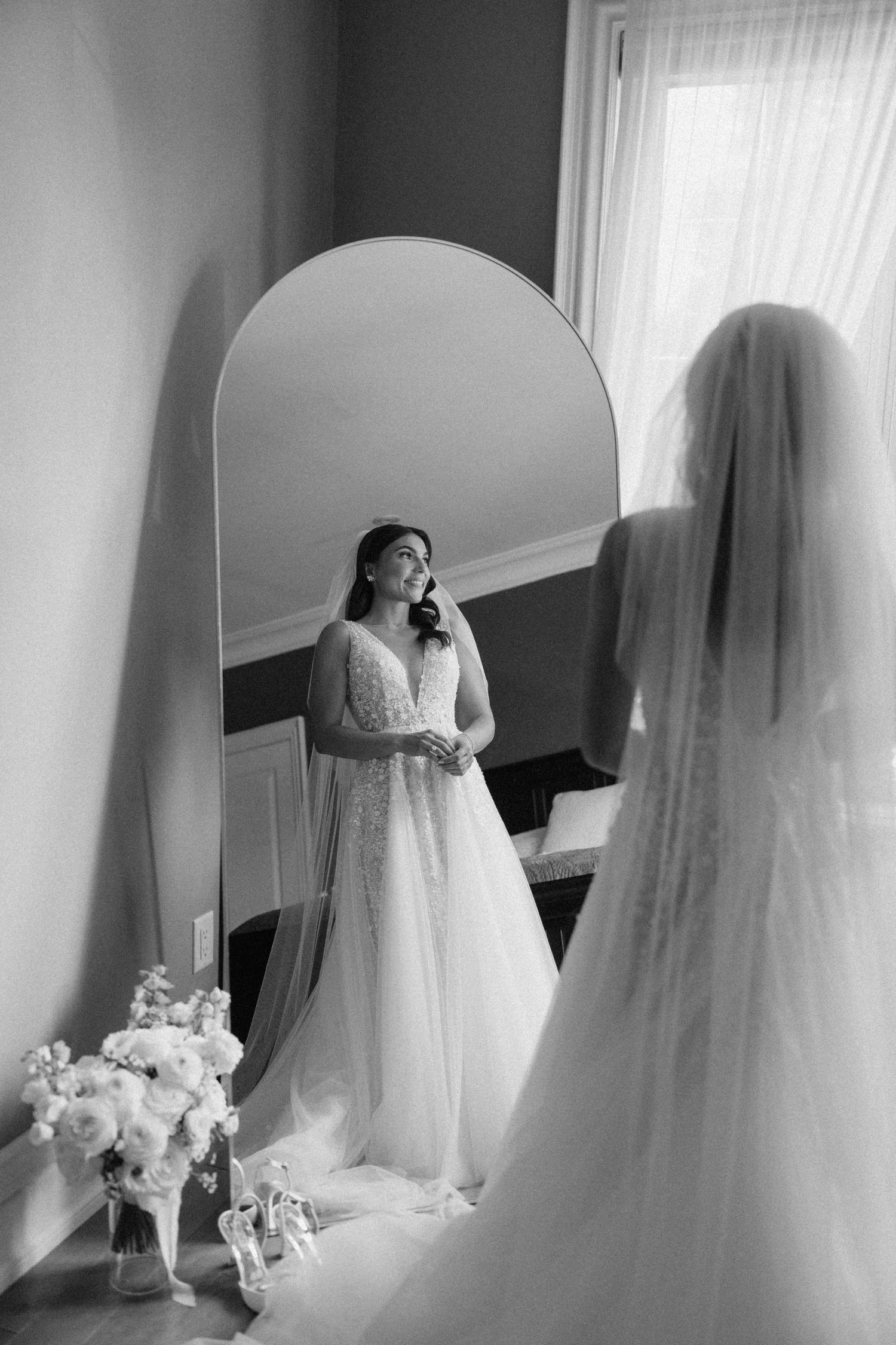 Bride in lace wedding gown and veil admires her reflection in an arched floor mirror in black and white.