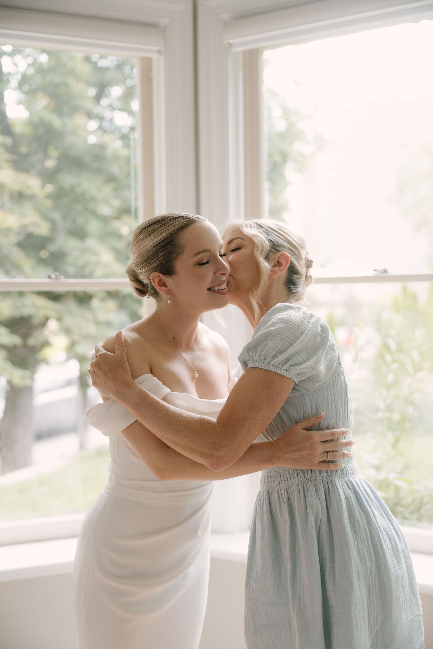 Bride and bridesmaid sharing a warm embrace and kiss near sunlit window.