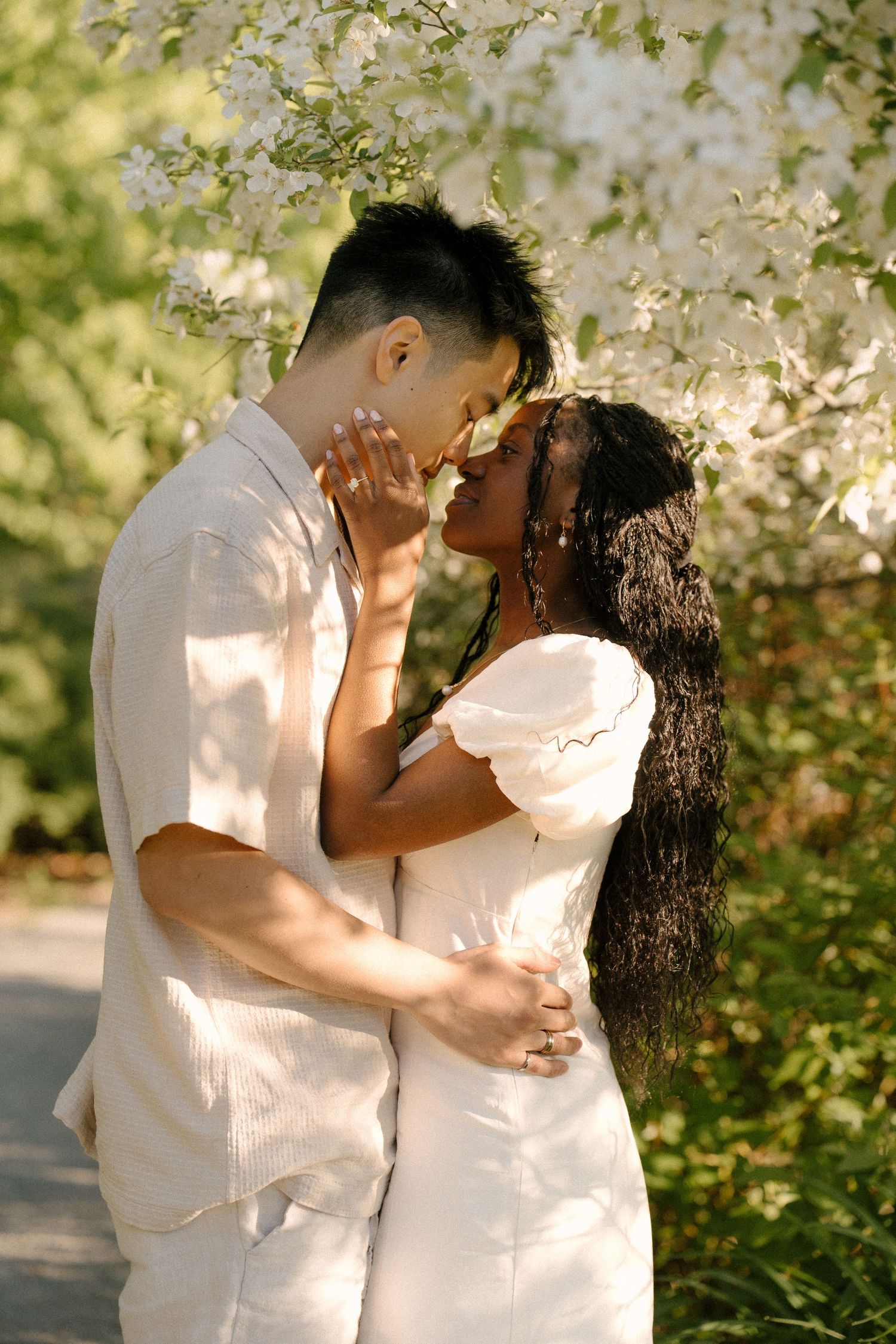 Interracial couple sharing a tender kiss under blooming white flowers in a sunlit garden, dressed in white outfits.