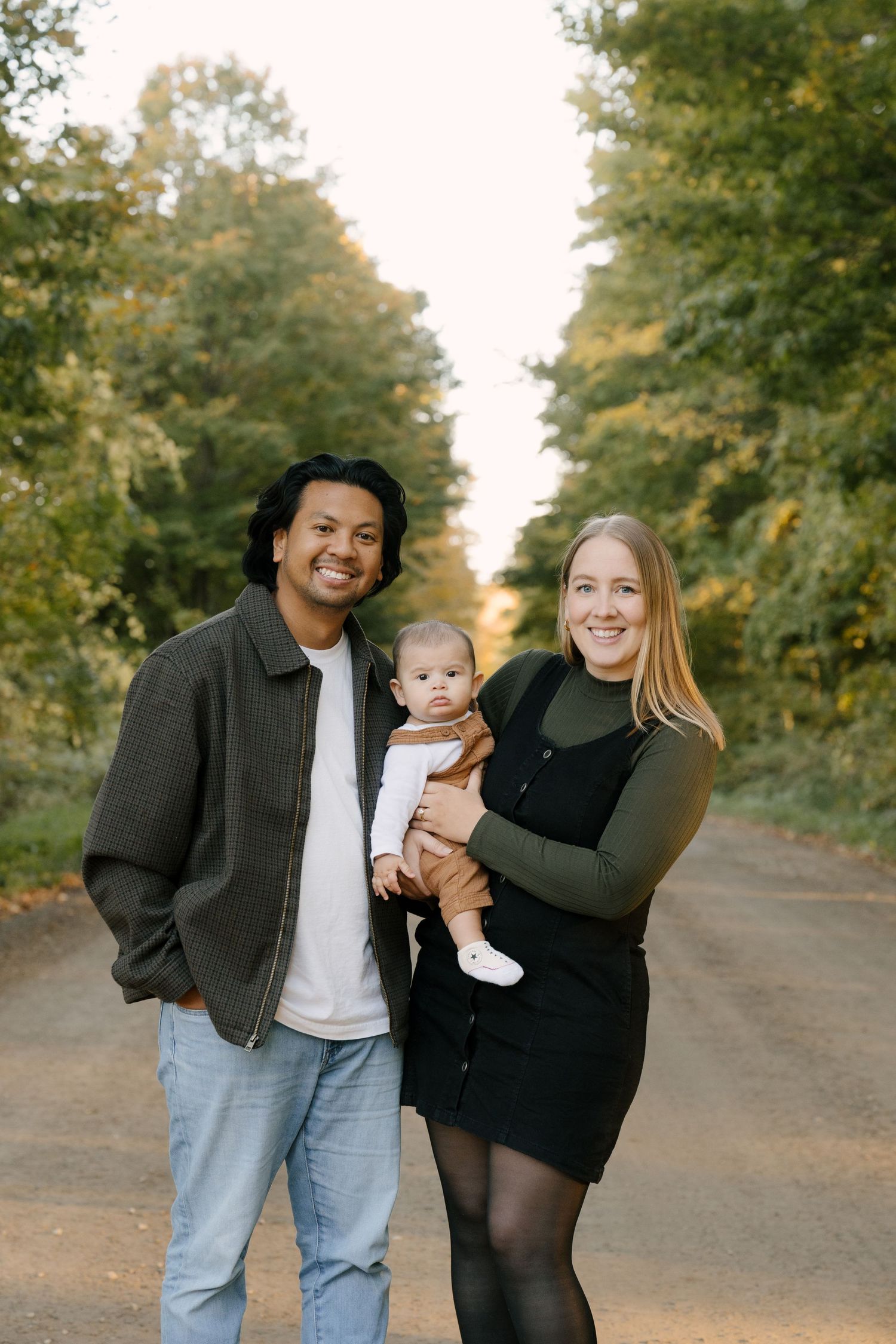 Smiling interracial family standing on tree-lined dirt road, mother holding baby in fall.