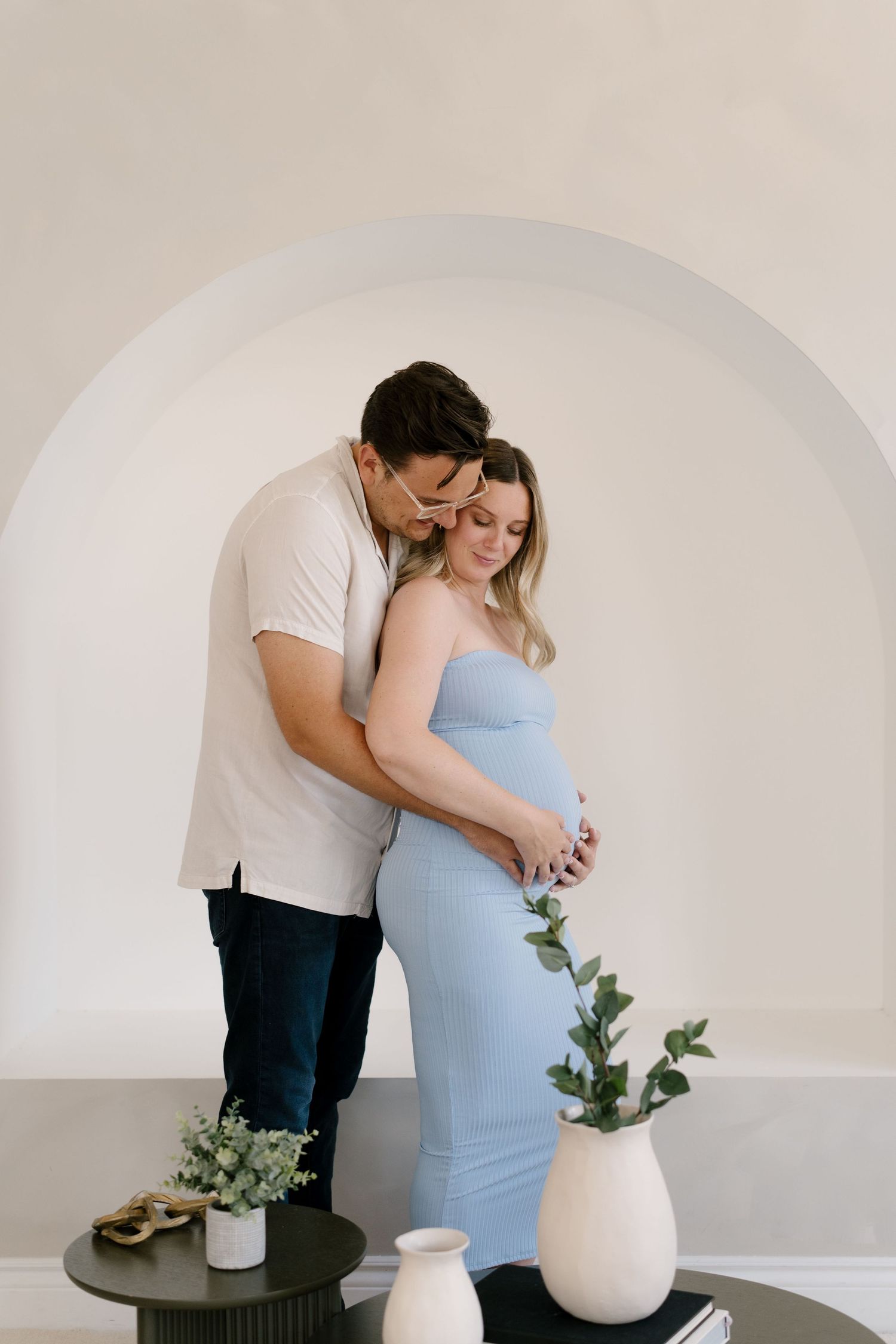 Man embracing pregnant woman in blue dress from behind under a white arch with decorative vases nearby.