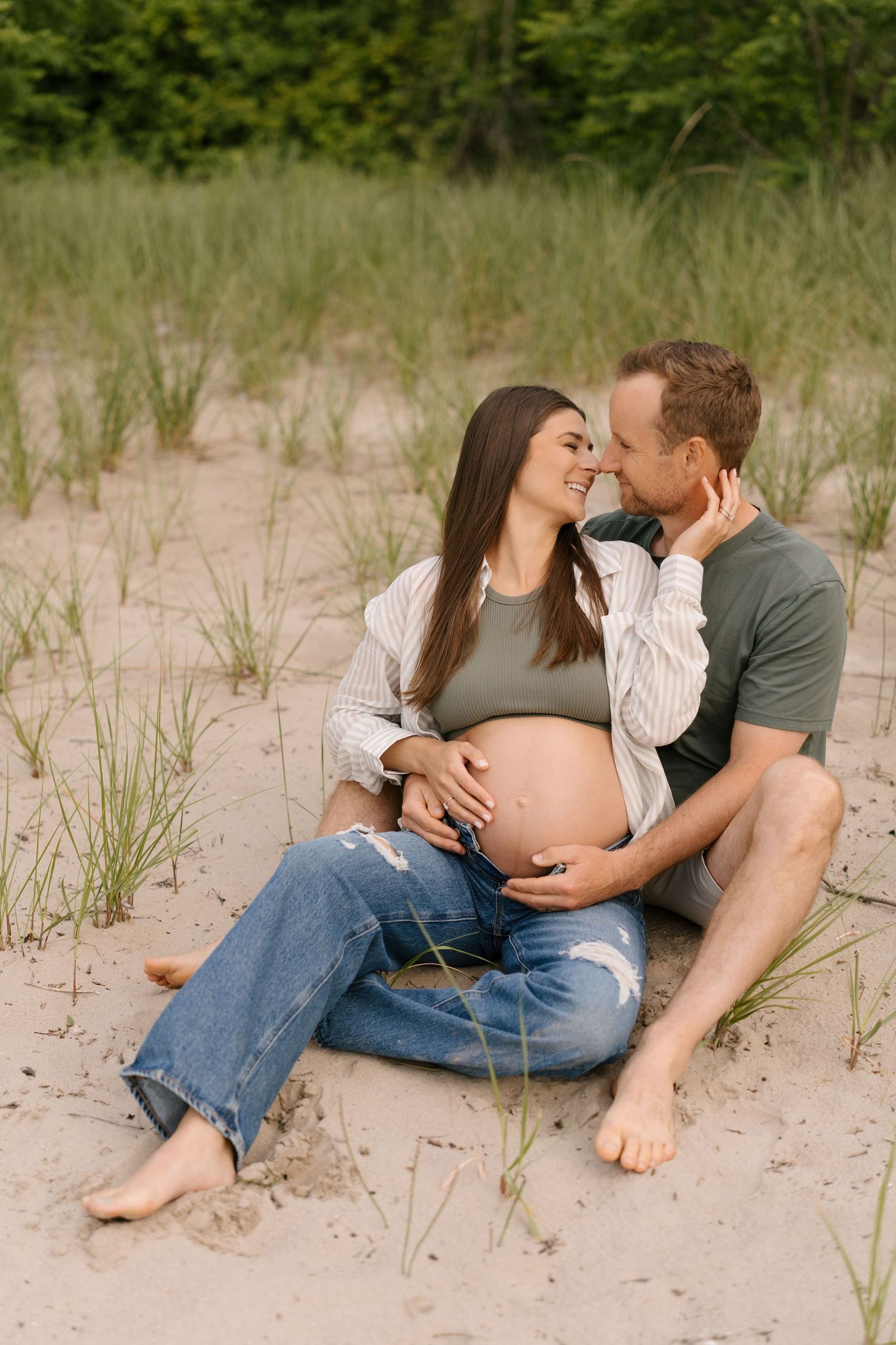 Couple sitting on sandy beach dunes, man tenderly holds pregnant womans baby bump as they share an intimate moment.