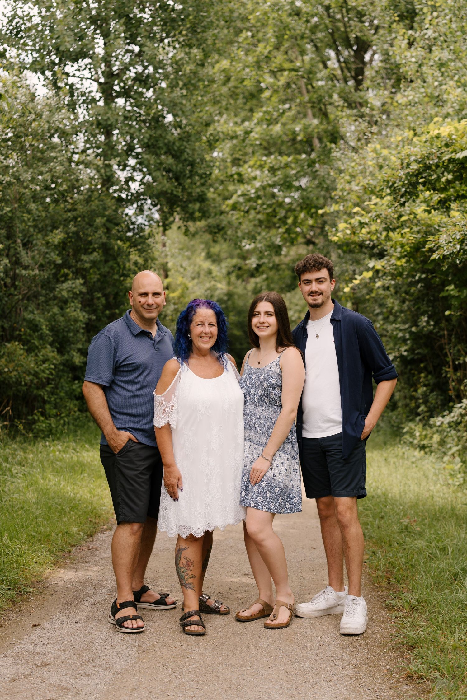 Family of four smiling together on a tree-lined path outdoors during summer, wearing casual attire.