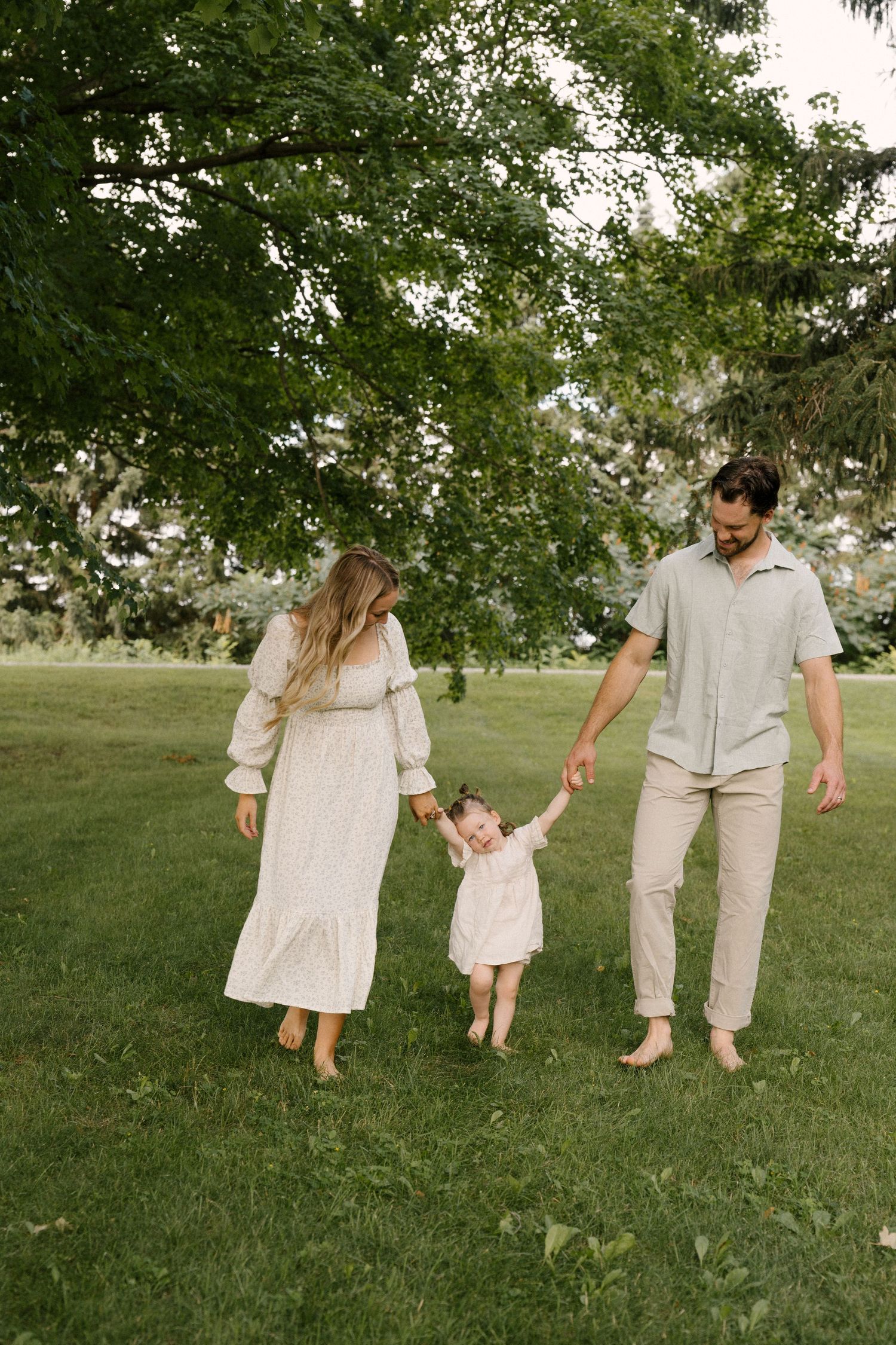 Parents swinging toddler by hands while walking barefoot on lush green grass outdoors.