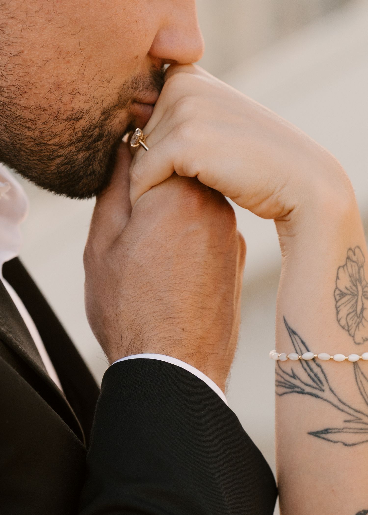 Groom kissing bride's hand showing engagement ring, with floral tattoo visible on her arm.