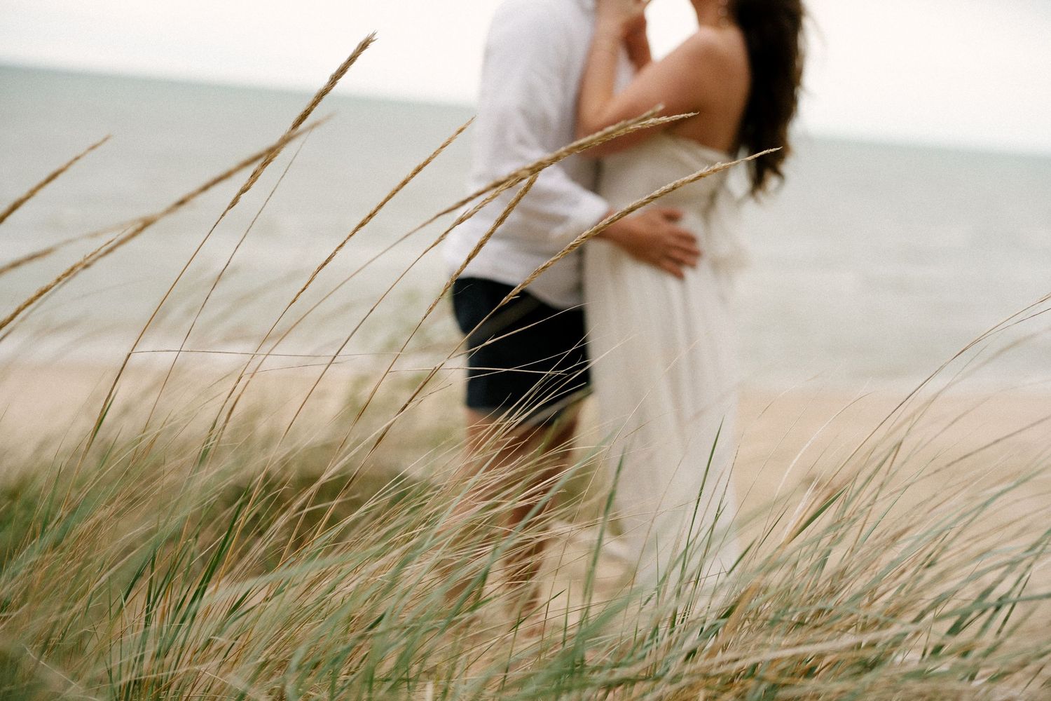 Couple embracing tenderly in tall golden beach grass with the calm ocean visible behind them.