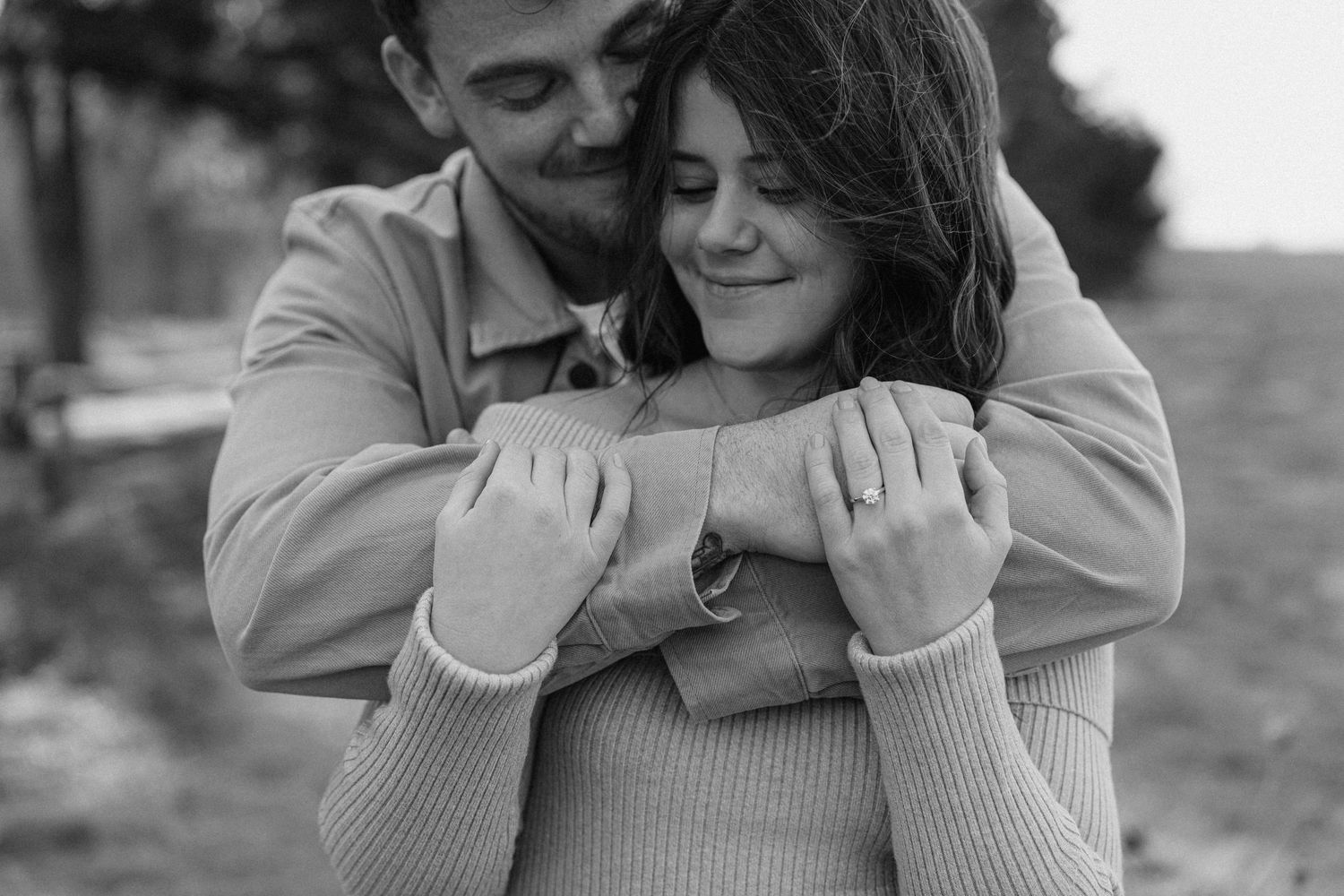 Black and white photo of man embracing woman from behind, both smiling warmly outdoors.