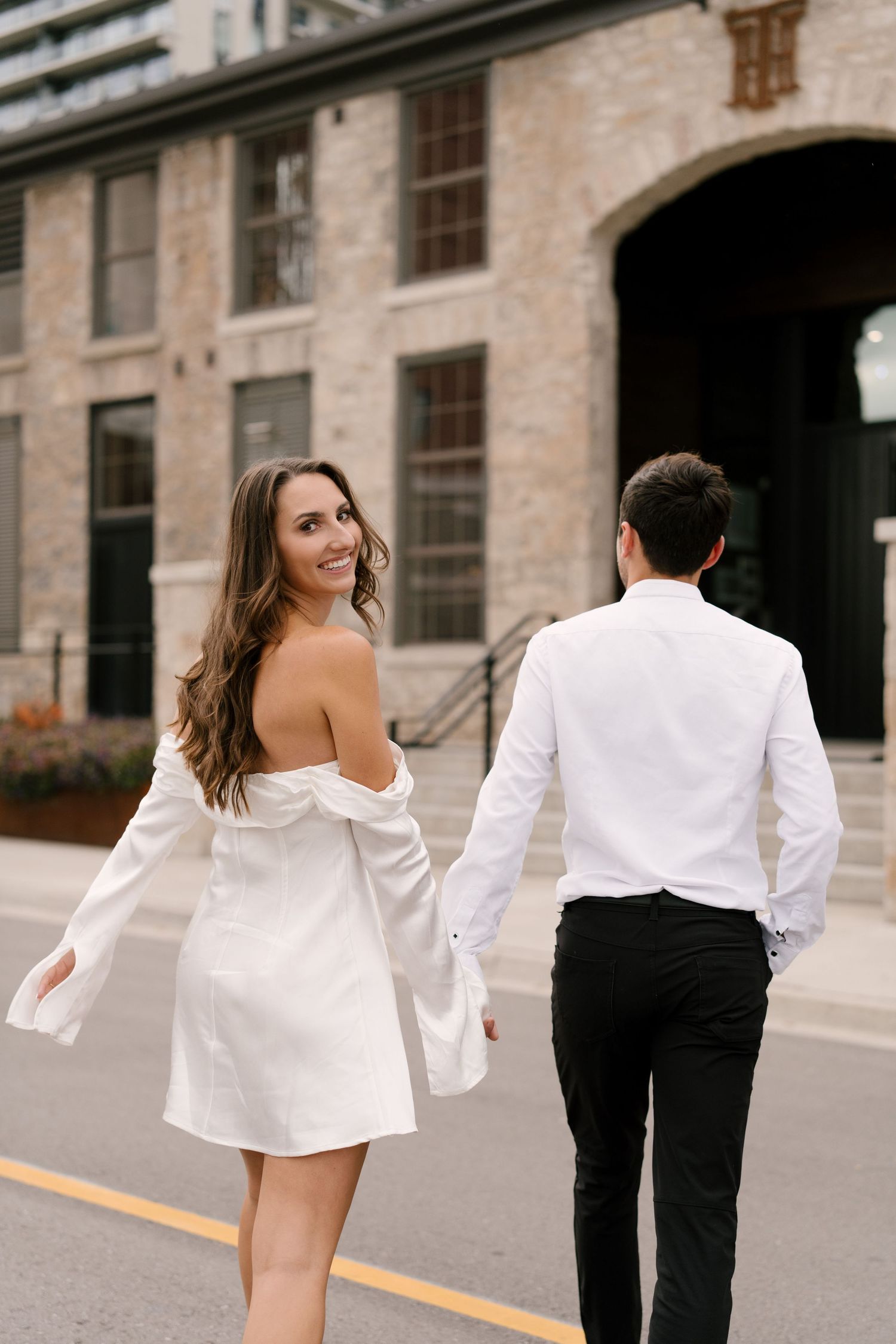 Couple walking on a city street, woman in white off-shoulder mini dress looks back smiling at camera.