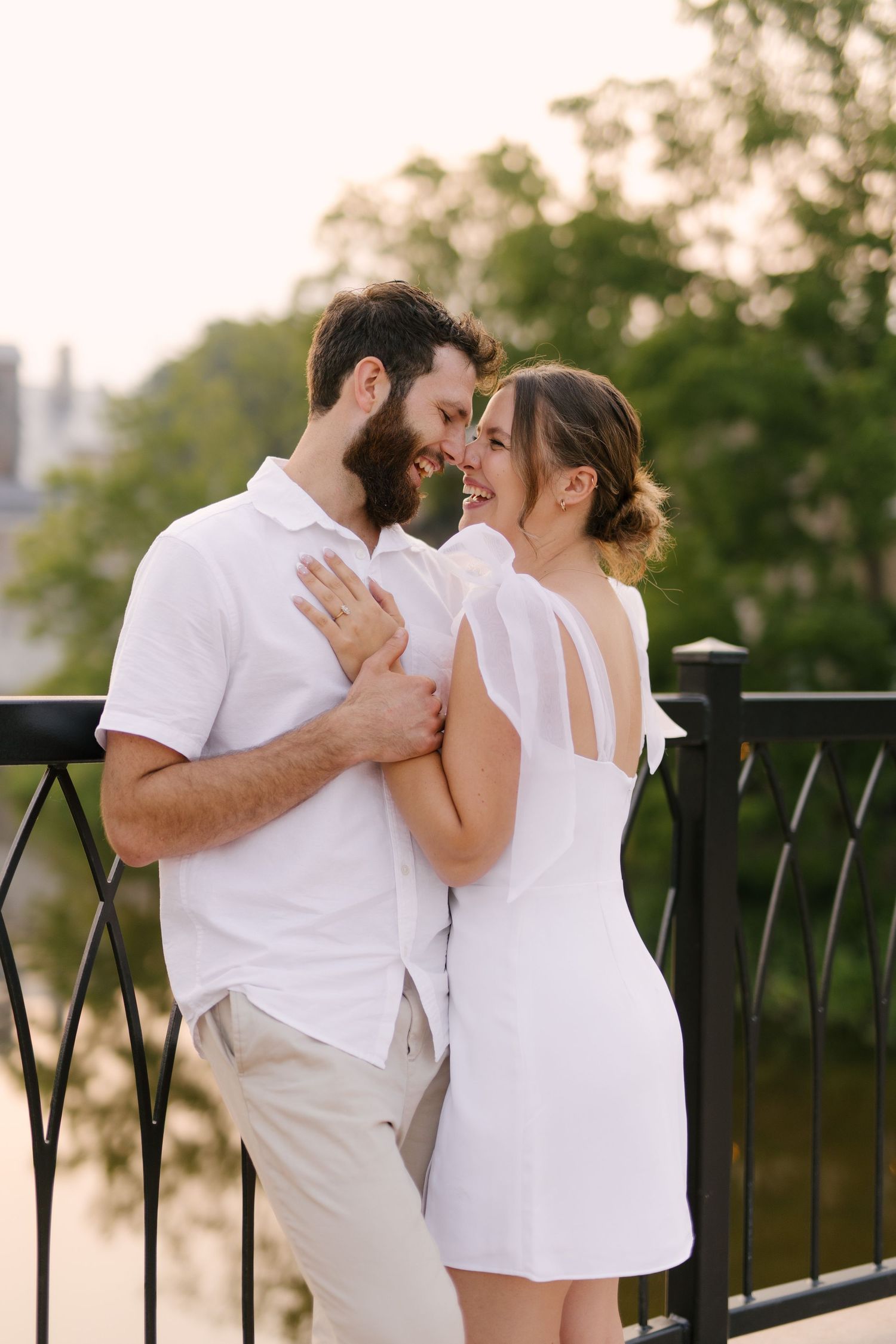 Loving couple in white outfits sharing an intimate moment on a bridge with lush green trees behind them.
