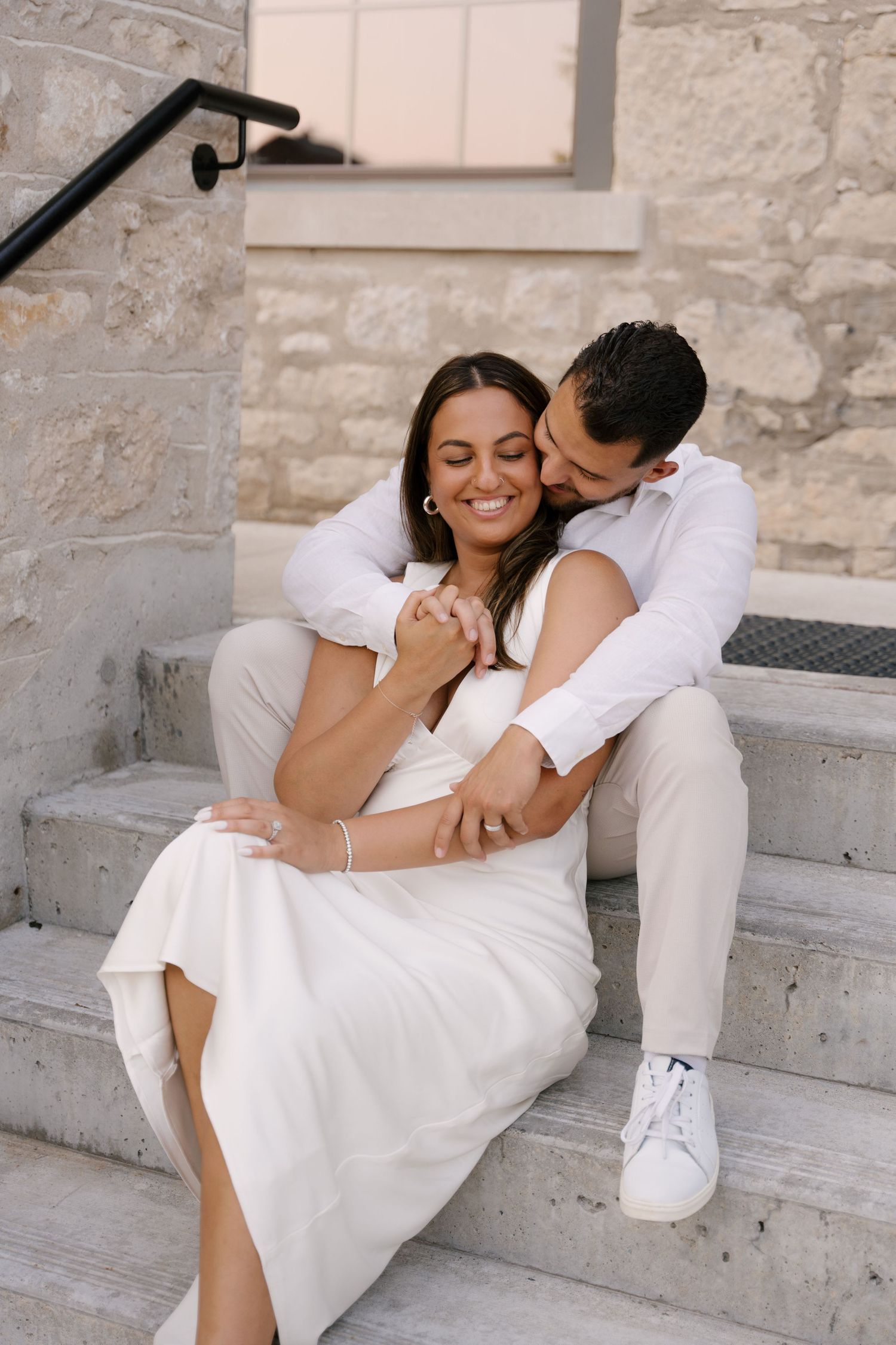 Romantic couple sitting on stone steps, man embracing woman in white dress at sunset.