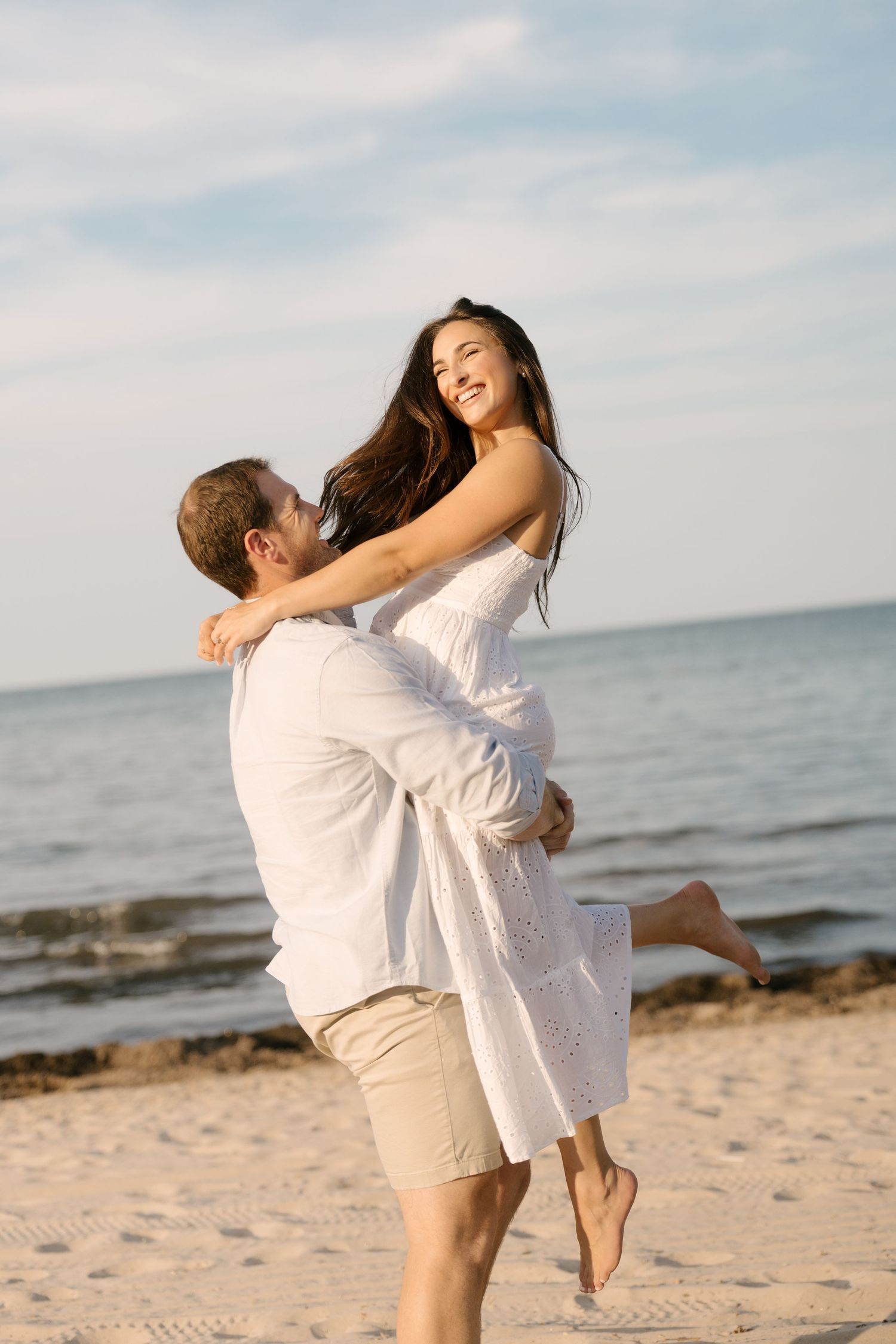 Man lifting smiling woman in white dress on sunny beach with ocean waves in the background.