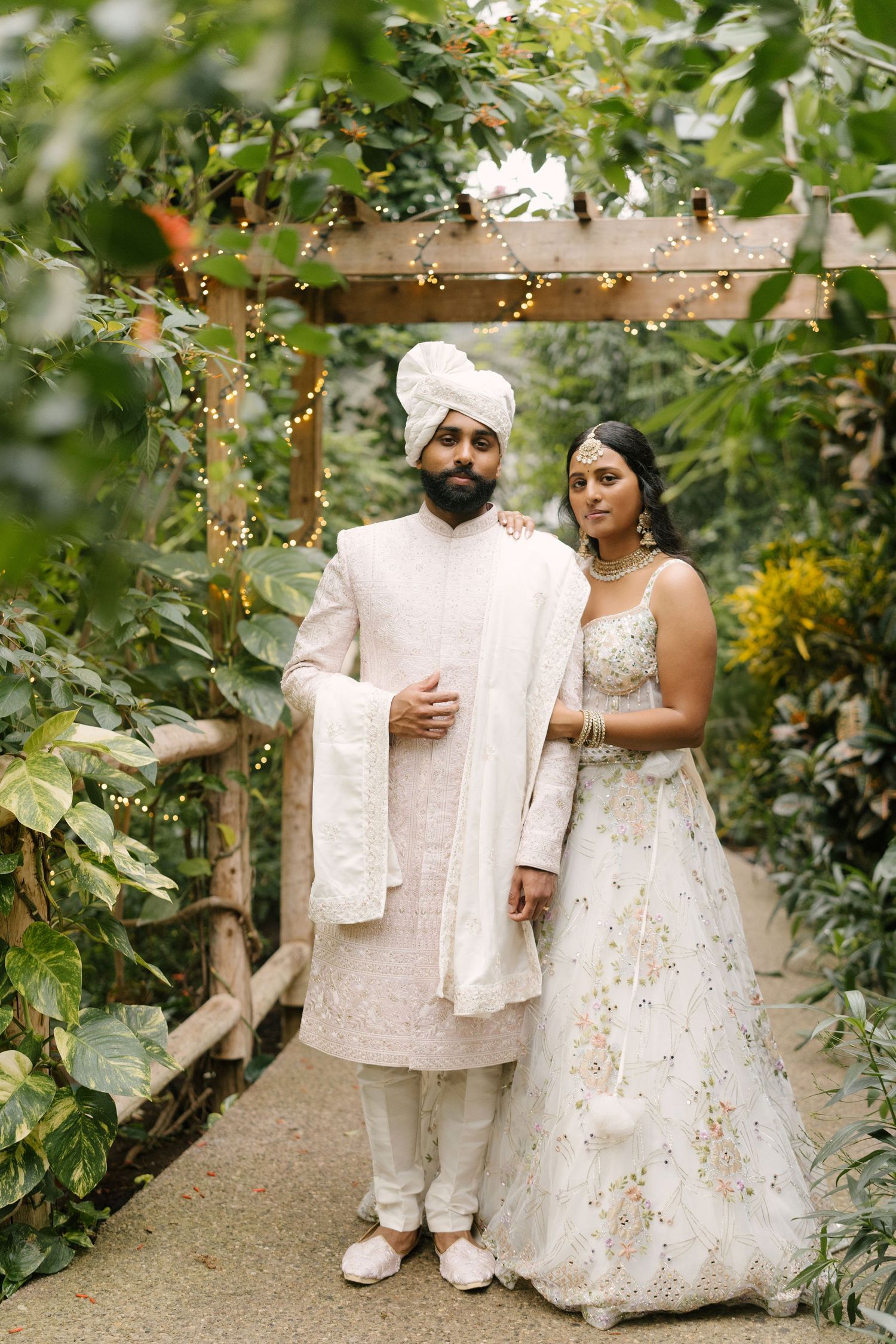 Indian bride and groom in matching ivory wedding attire pose under leafy garden pergola.