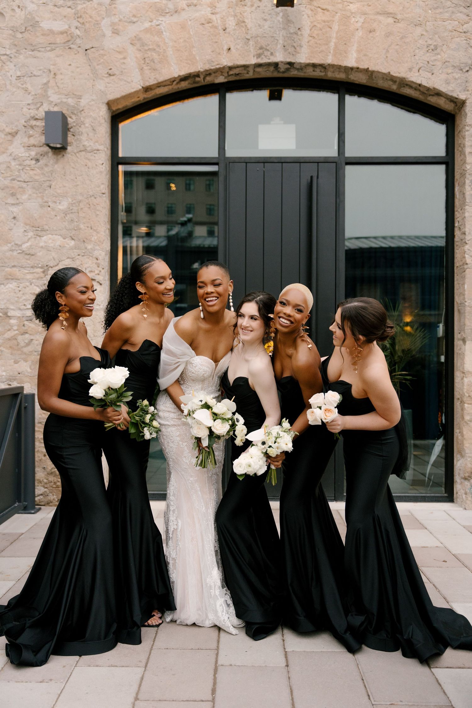 Bride in white lace gown poses with five bridesmaids in black strapless mermaid dresses holding white bouquets.