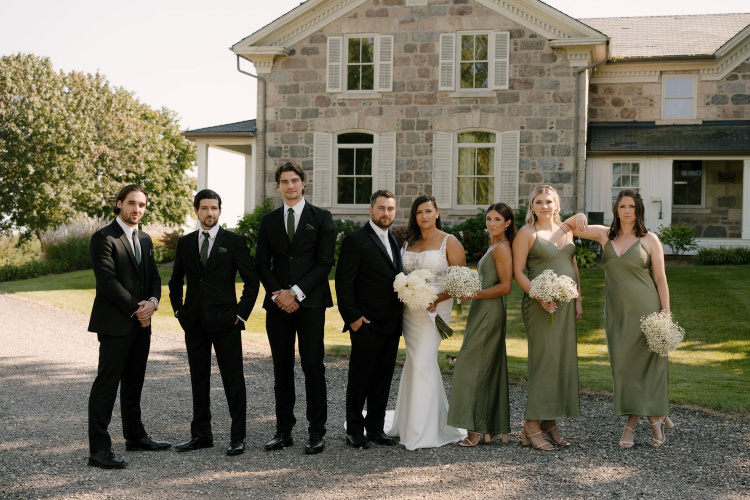 Wedding party poses in front of stone Wakefield Estates with groomsmen in black and bridesmaids in sage.