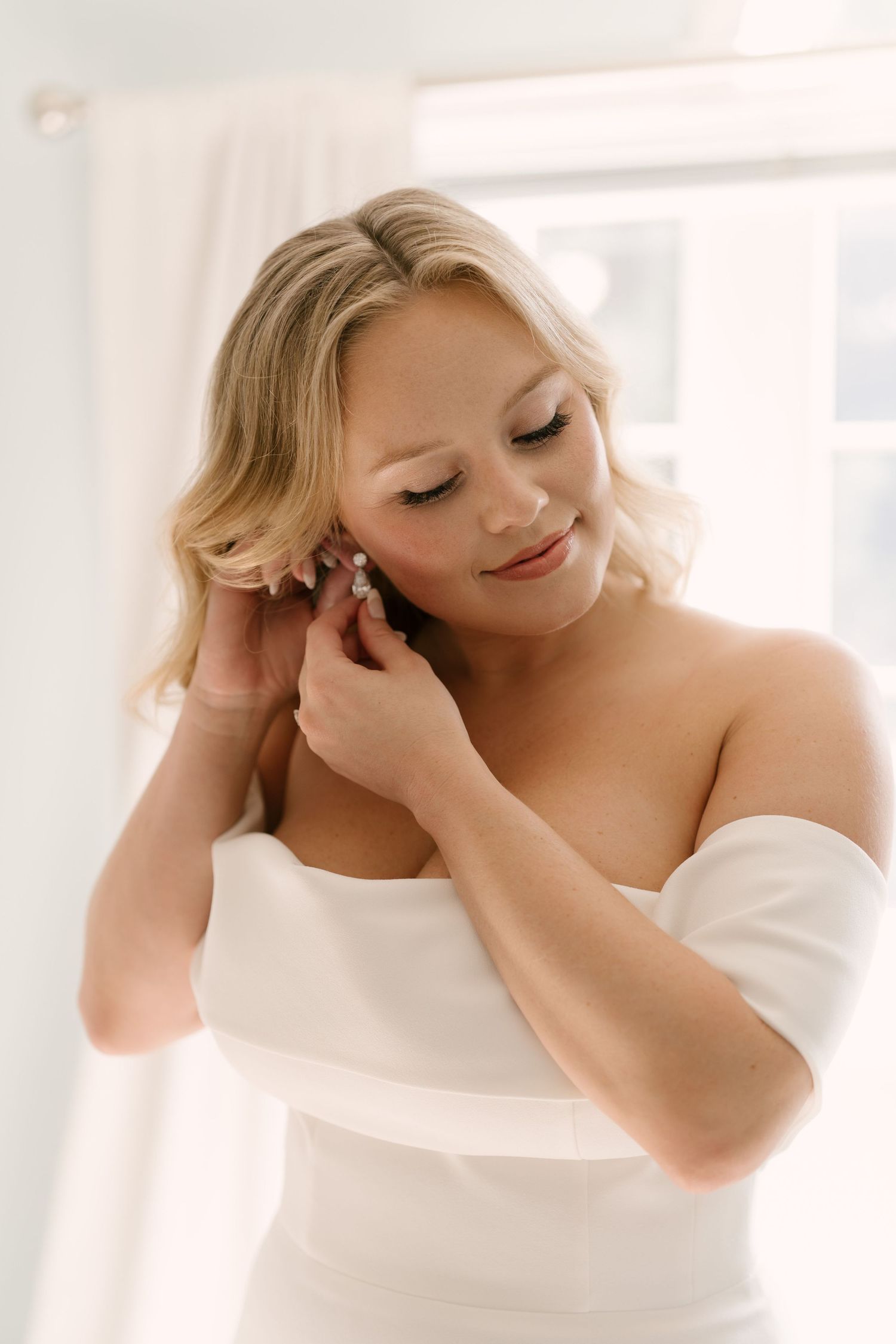 Blonde bride in off-shoulder white gown puts on earrings near bright window while getting ready.