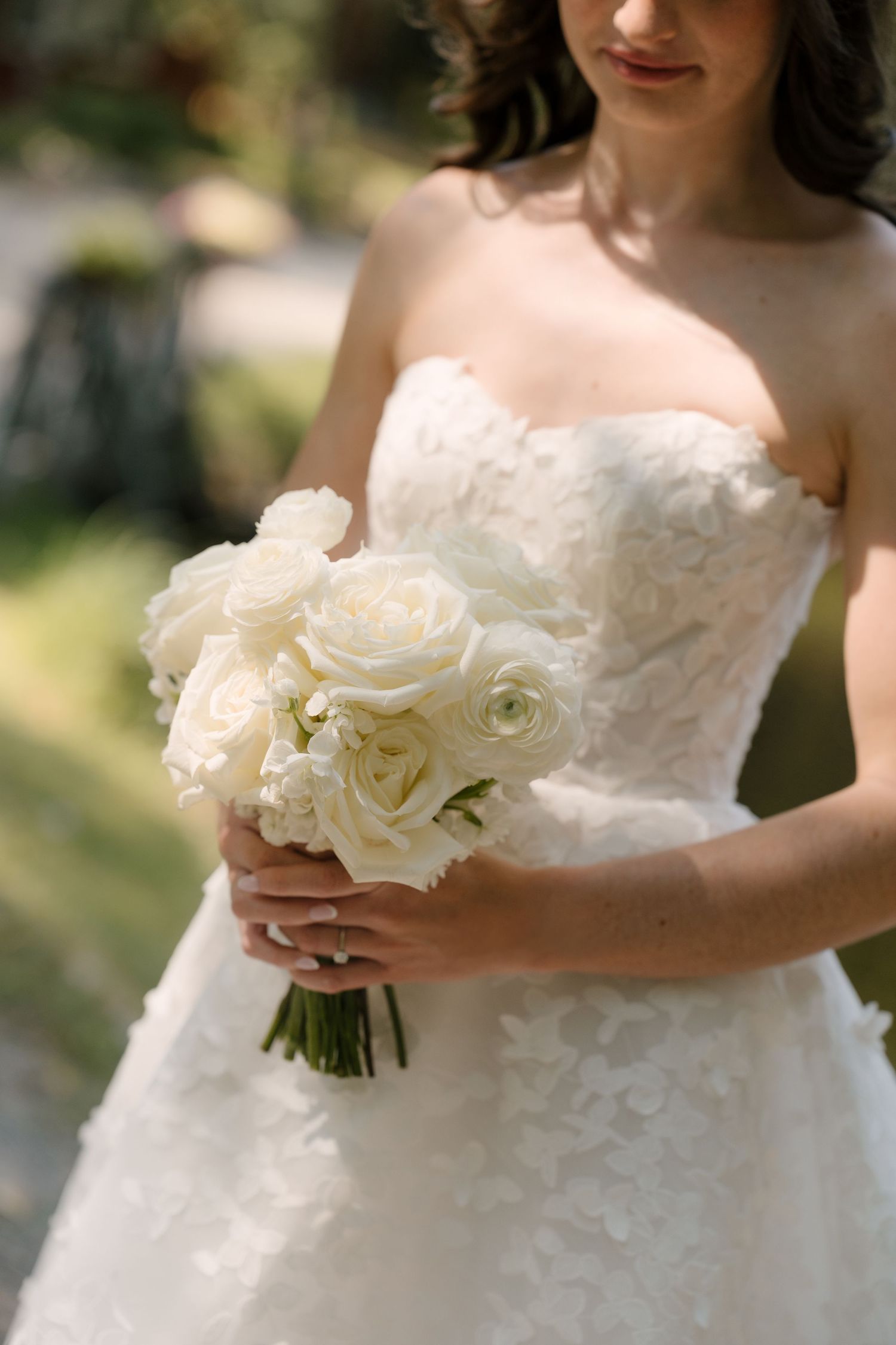 Bride in strapless white floral gown holding elegant all-white rose bouquet outdoors on her wedding day.