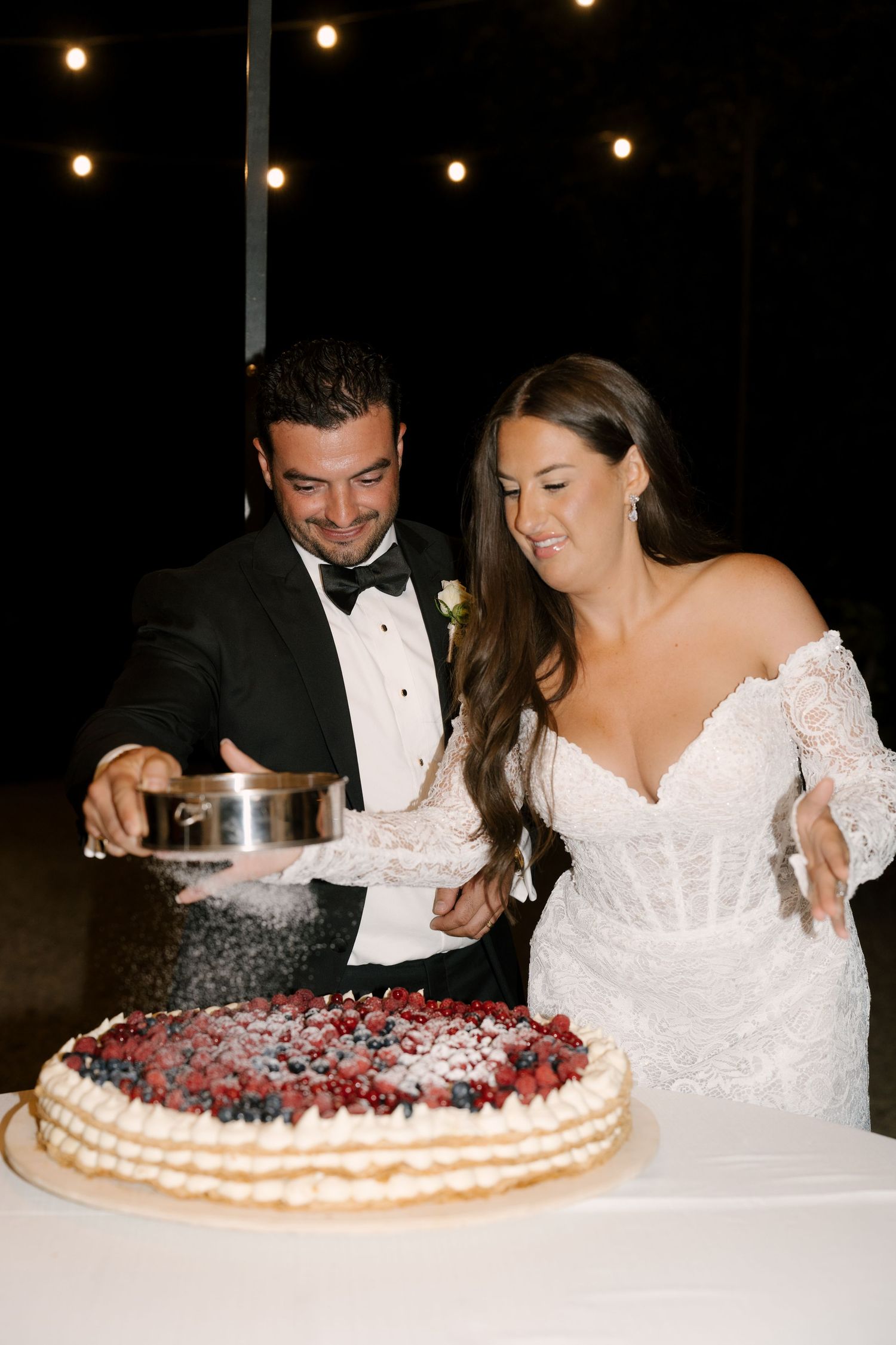 Bride and groom cutting berry-topped wedding cake together outdoors at night celebration.