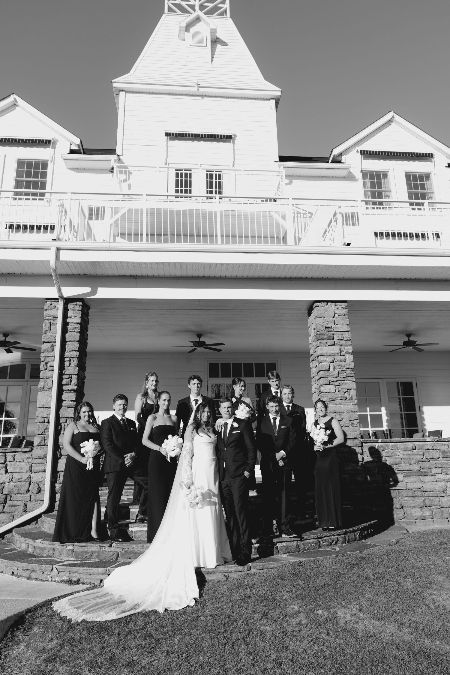 Black and white formal portrait of bride, groom, and full wedding party posed outside elegant venue.