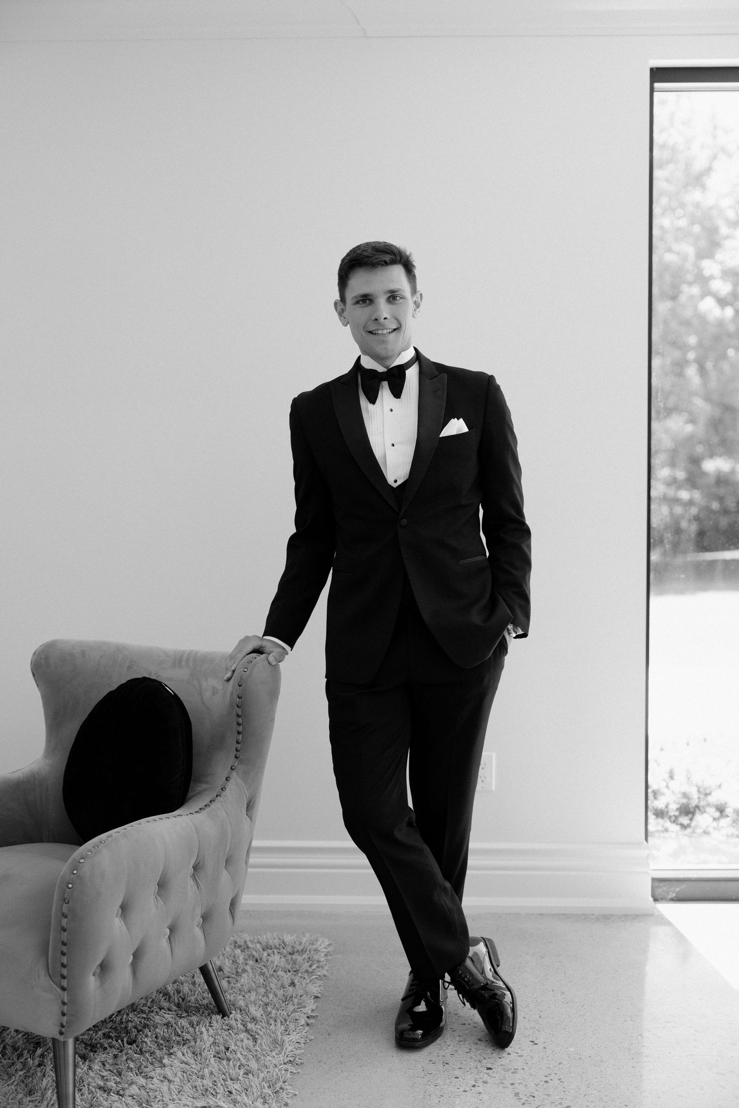 Dapper groom in black tuxedo leaning against tufted chair in bright modern room, black and white photo at Roseville Estates wedding venue.