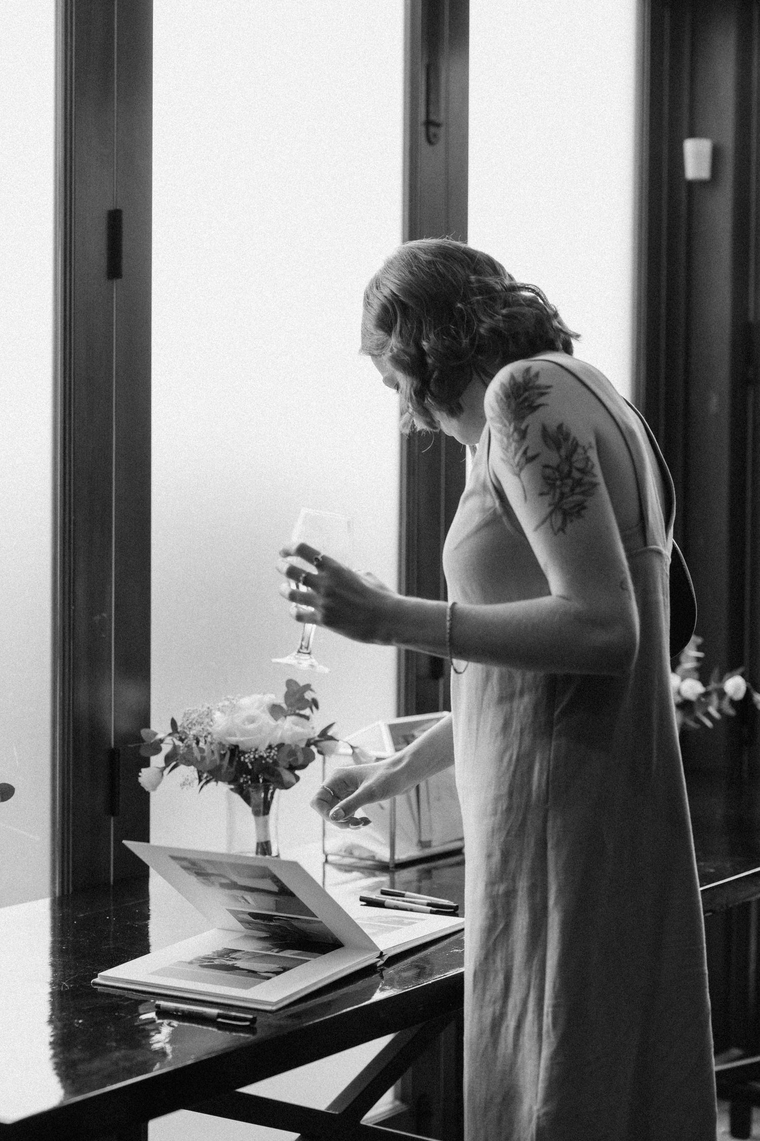 Woman in elegant dress holds champagne glass while reviewing wedding materials by sunlit window.