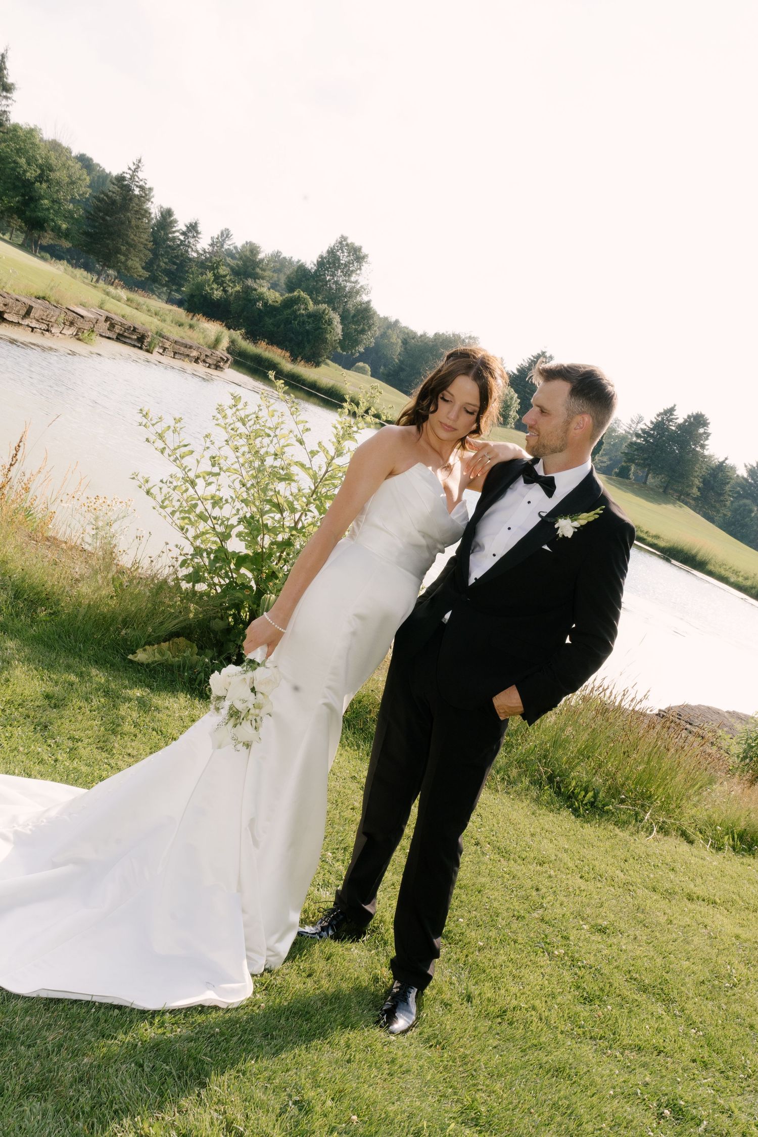 Bride in white gown and groom in black tuxedo share a romantic moment on green lawn beside a serene pond.