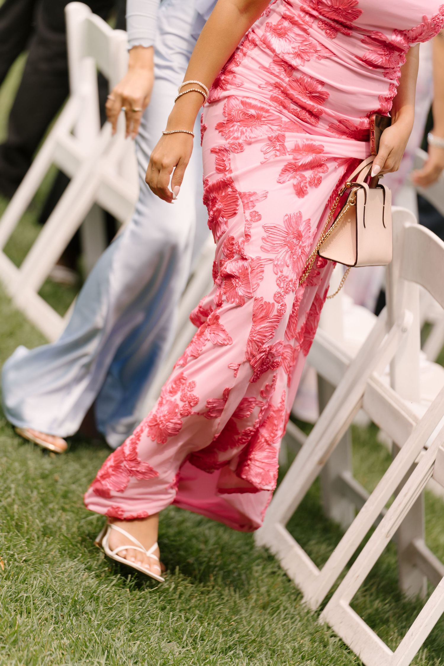 Woman in pink floral maxi dress holding a beige handbag walks between white chairs on green grass.