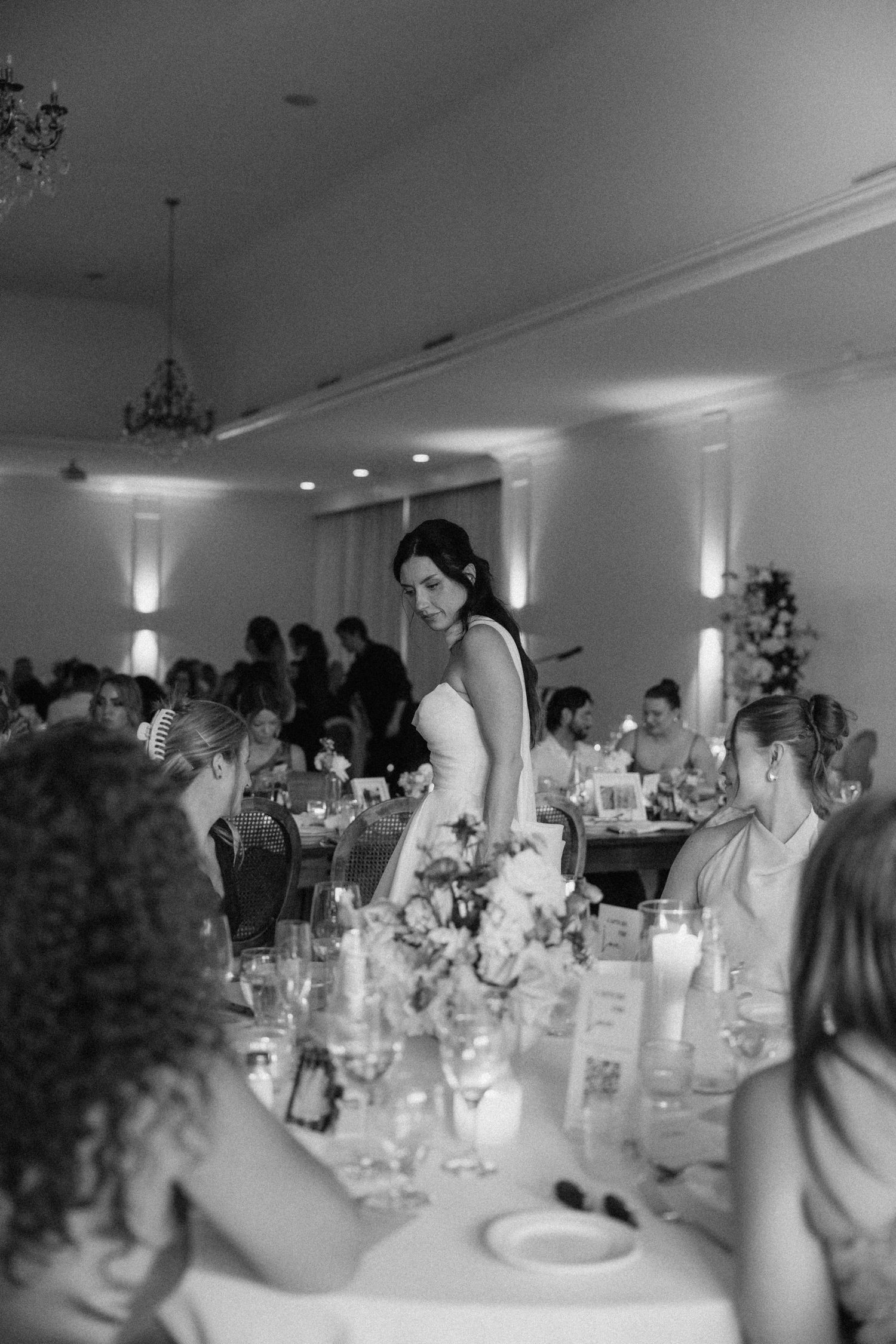 Black and white photo of bride standing among guests at elegantly decorated wedding reception dinner tables.