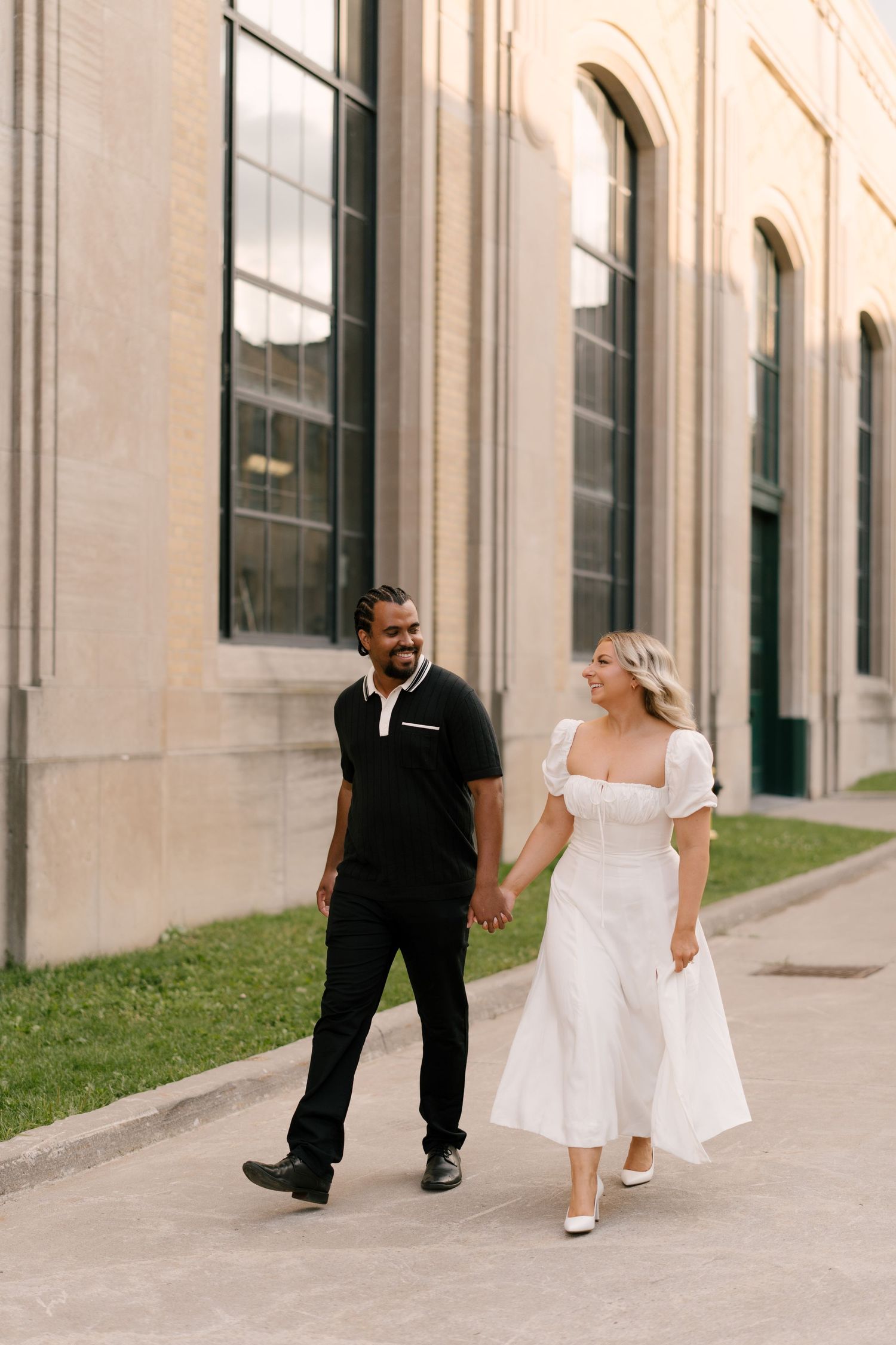 Interracial couple walking hand in hand past stone building, woman in white puff-sleeve midi dress smiling.