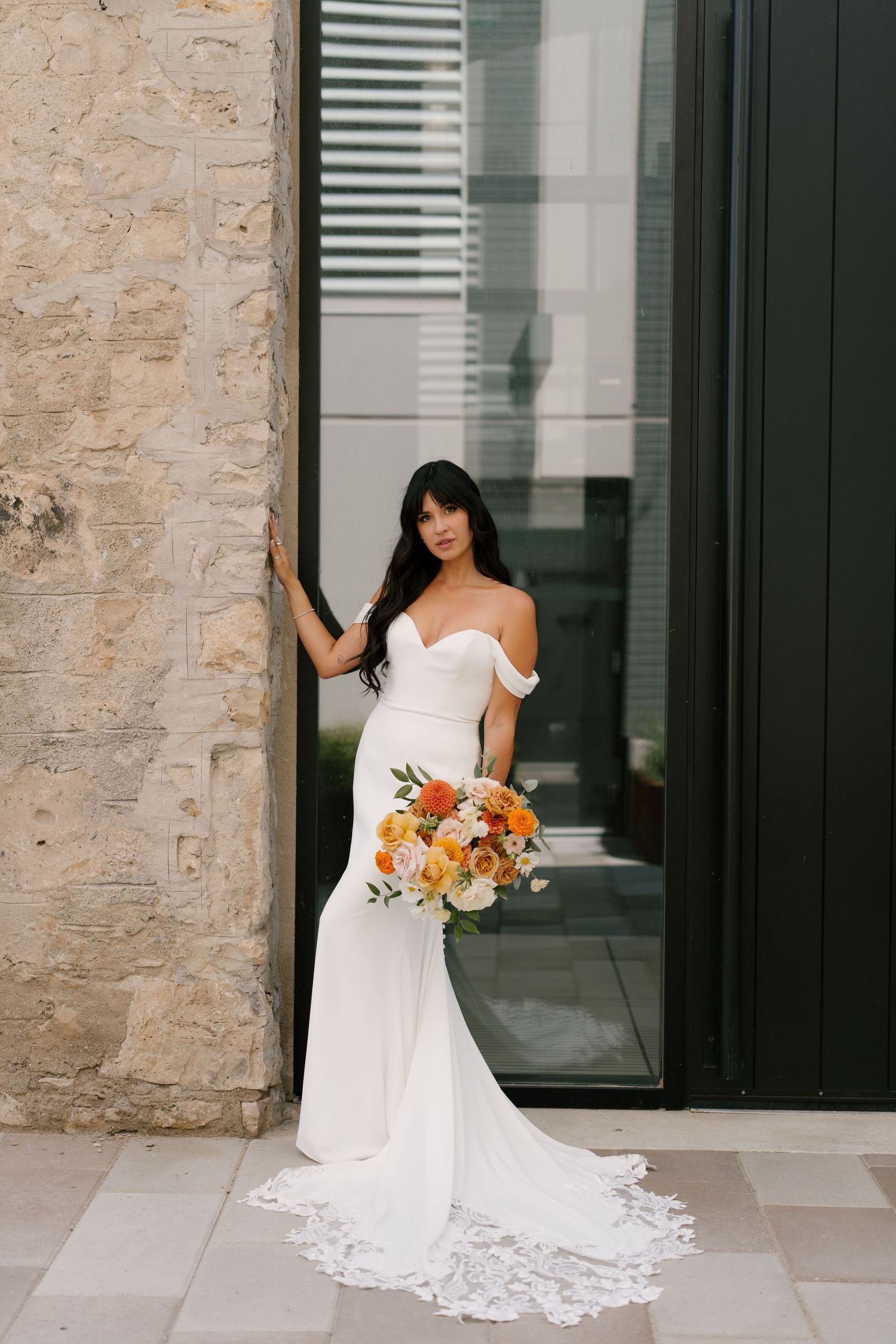 Elegant bride in off-shoulder white fitted gown with lace train holds vibrant orange and white bouquet by stone wall outside Tapestry Hall.