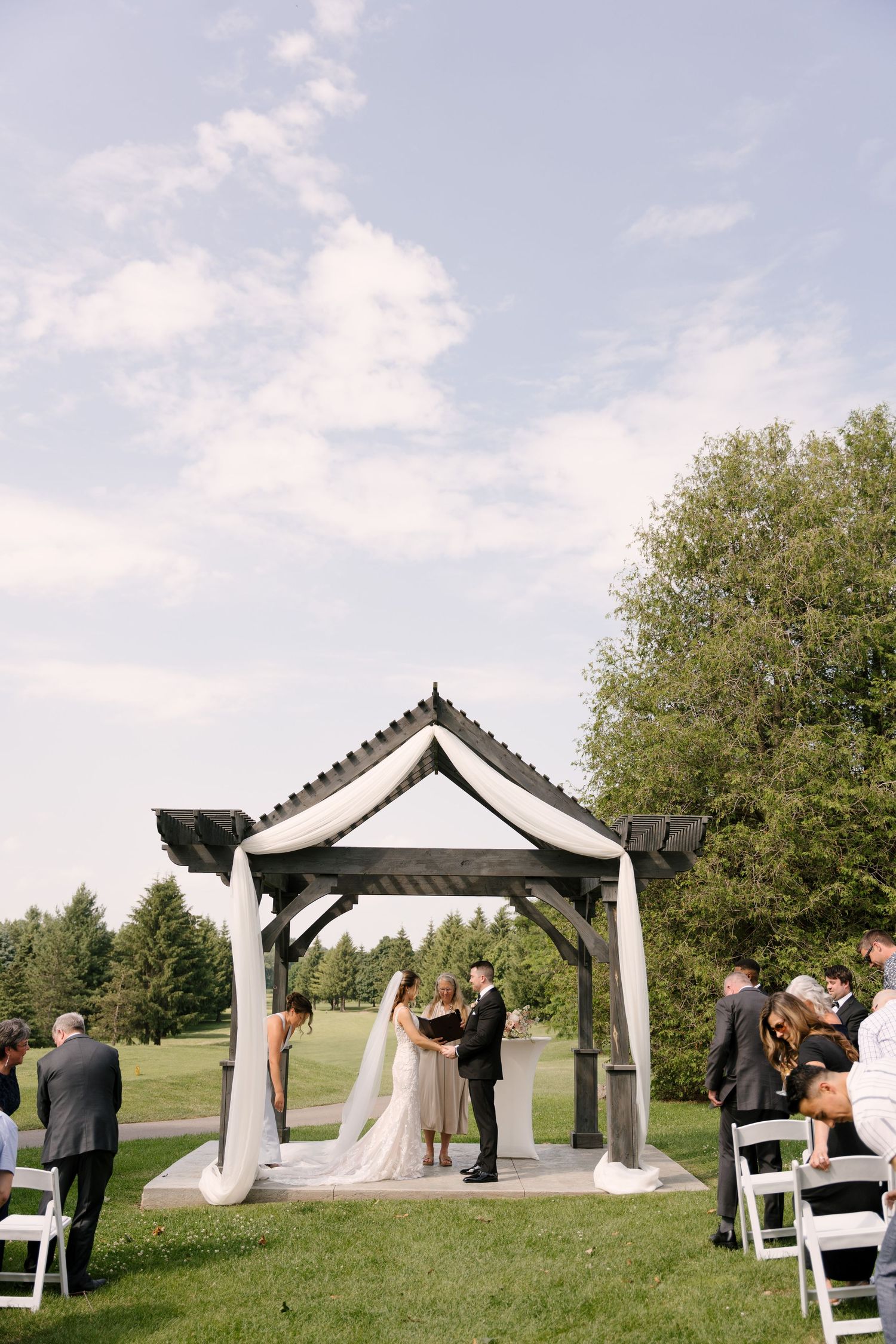 Outdoor wedding ceremony under a dark wood gazebo on a green lawn with guests seated in white chairs at St Marys Golf Club.
