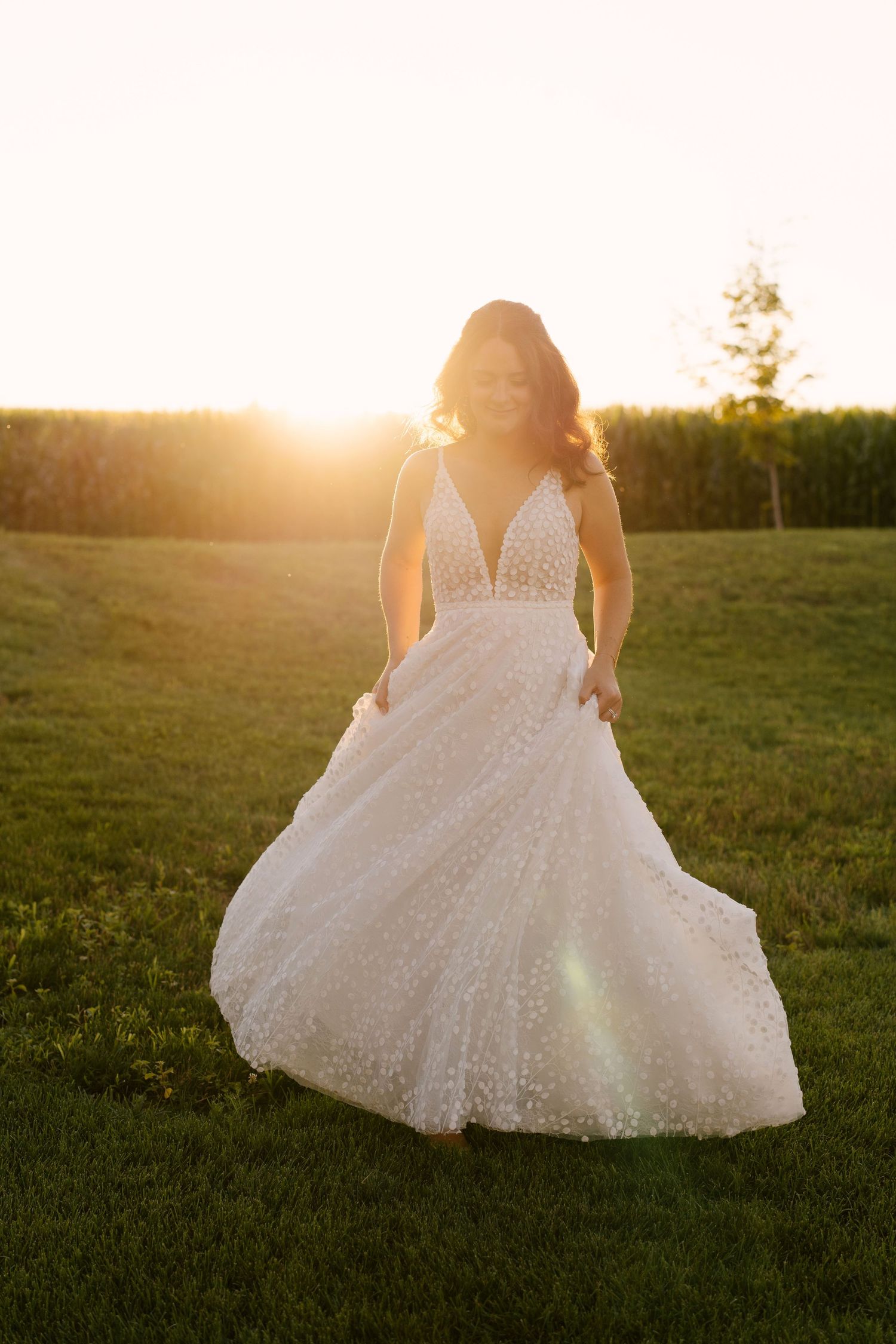 Bride twirling in embellished white wedding gown at golden sunset in an open green field.